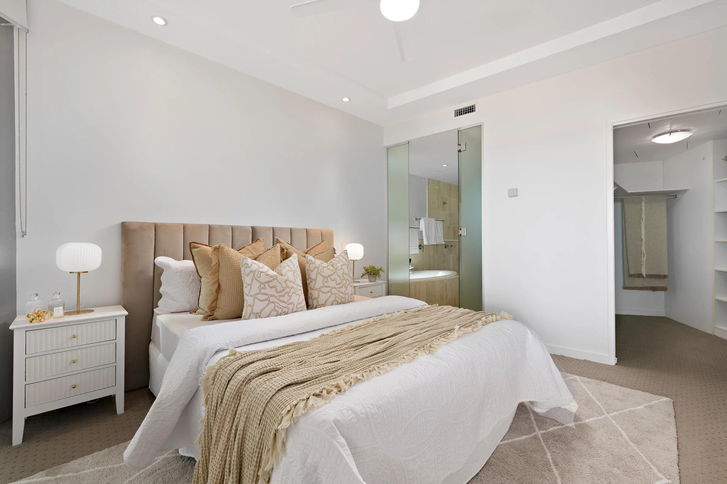 Modern bedroom with beige upholstered headboard, white bedding, and beige decorative pillows. Nightstand with white drawers, gold handles, and a white lamp. Open bathroom with a glass door showing a bathtub and towels. Ceiling with recessed lighting and a white fan. Carpeted floor with a beige and white geometric rug.