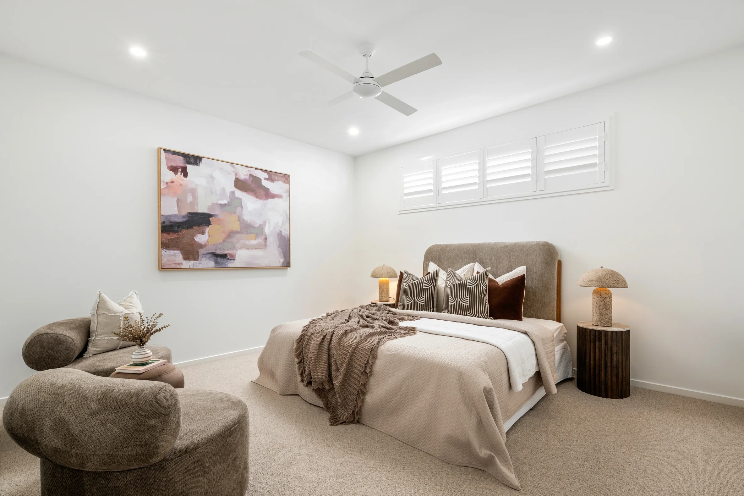 A cozy bedroom with a bed covered in neutral bedding, a brown throw blanket, and decorative pillows, flanked by matching table lamps on wood side tables, with abstract artwork on the white wall and a window with shutters.