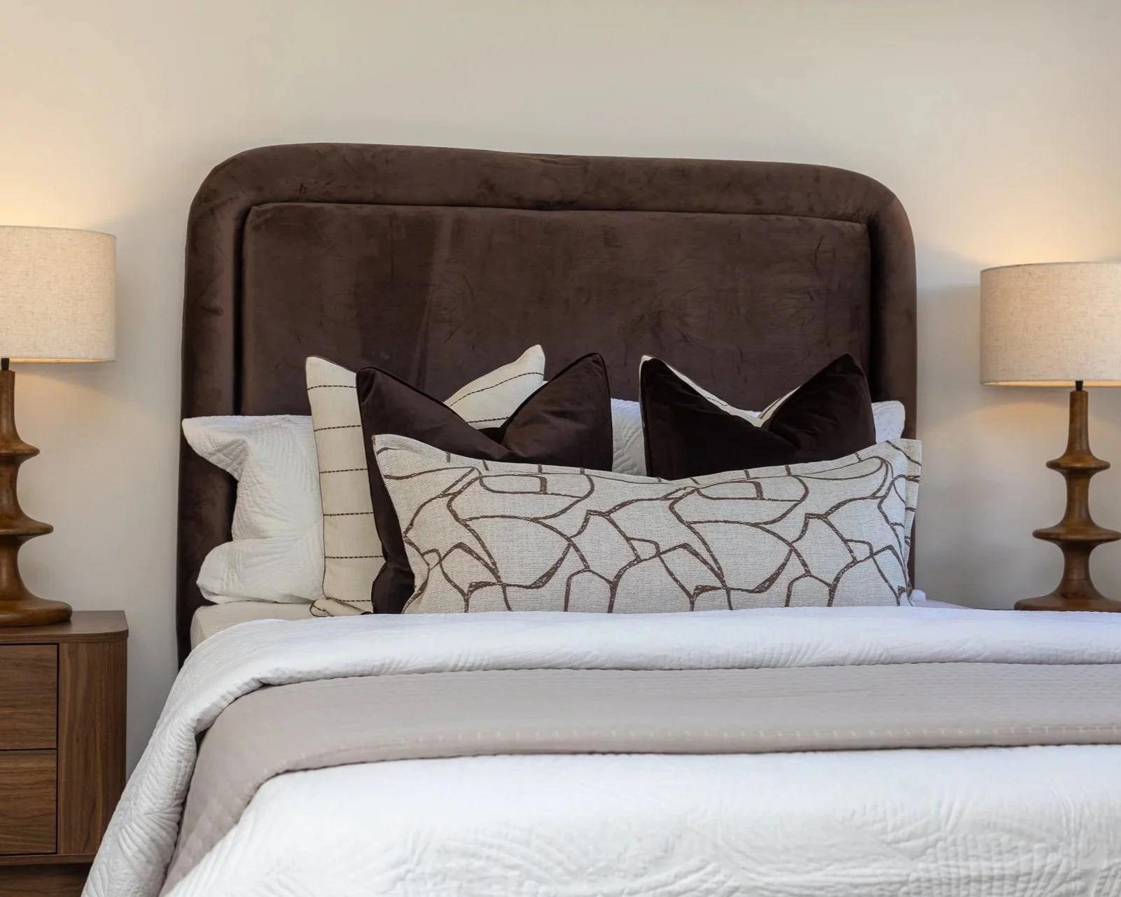 A neatly made bed with a tall, dark brown upholstered headboard, several decorative pillows in neutral tones with various patterns, and two wooden bedside tables with matching lamps on each side.