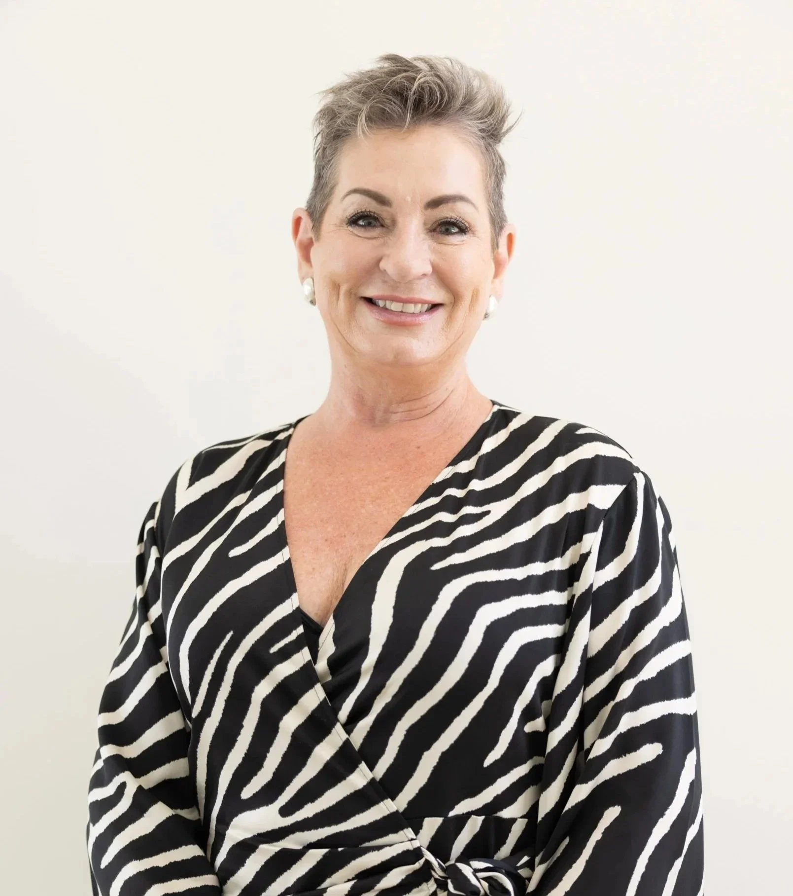 A smiling woman with short, styled gray hair, wearing pearl earrings and a black and white zebra print top, standing against a plain light background.