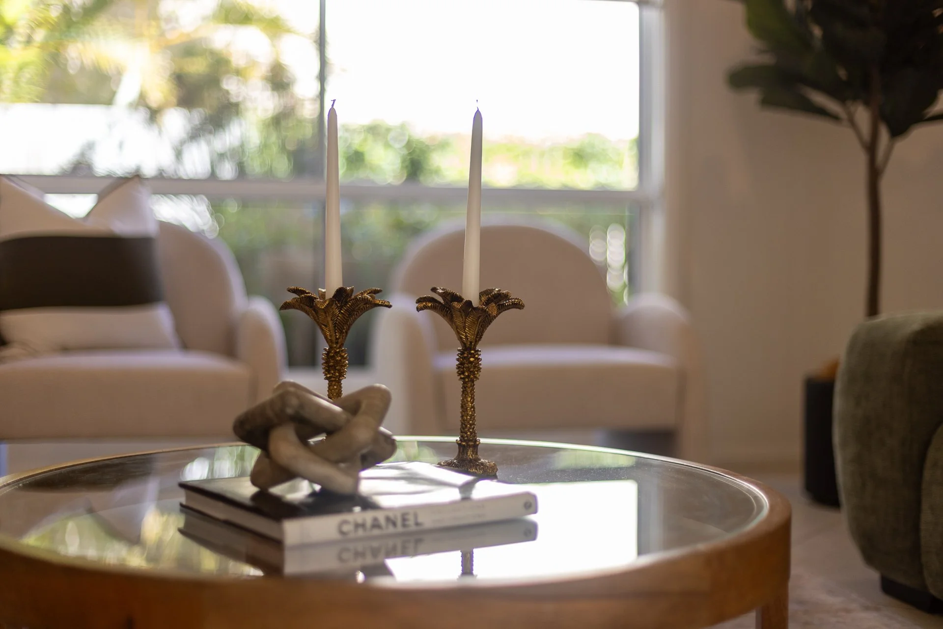 A living room with a glass-topped table holding decorative items, including a pair of ornate gold candle holders, a small coral sculpture, and a stack of books with 'Chanel' on the spine, in front of a window with natural light and a large potted plant to the side.