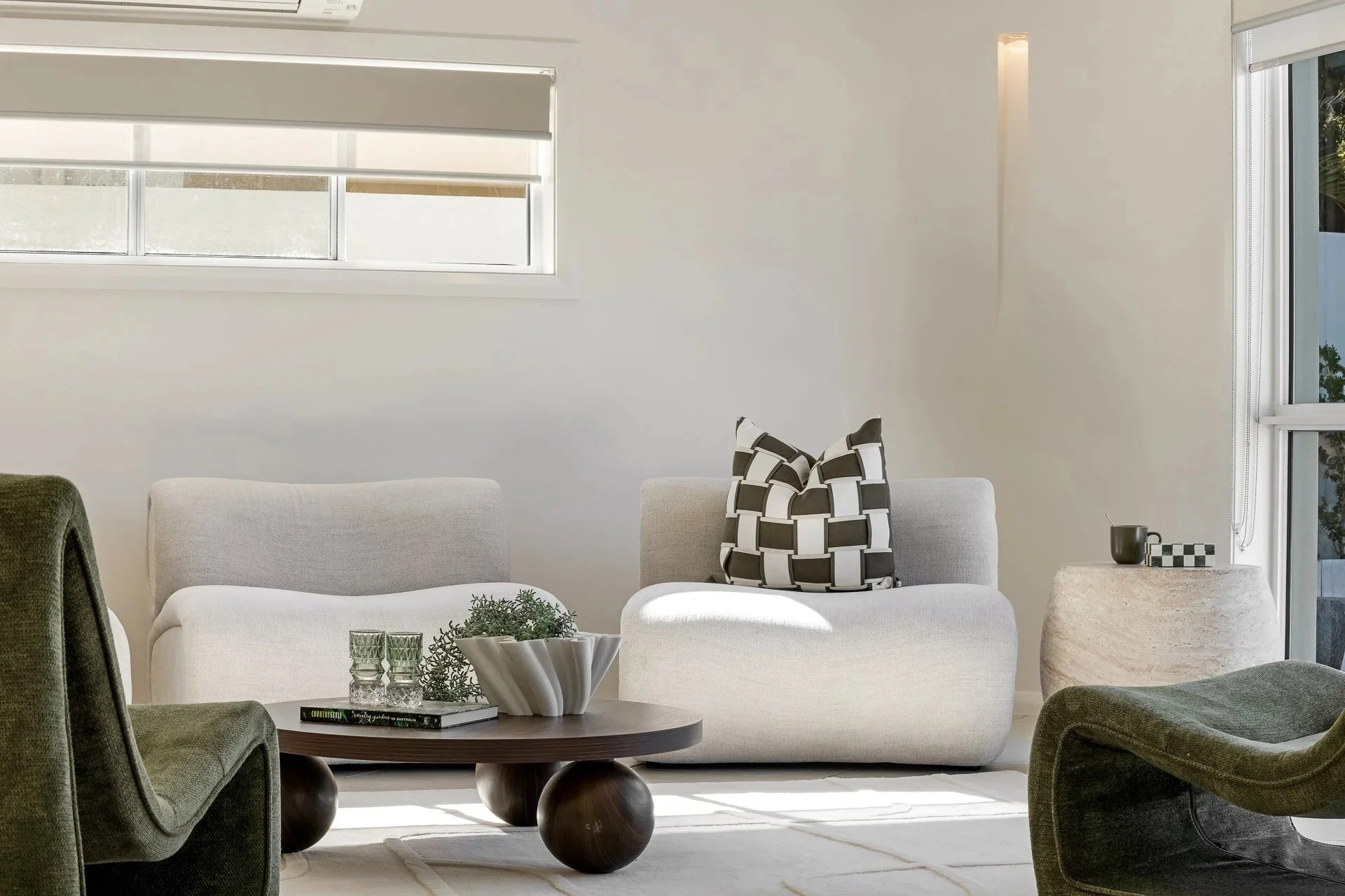 Modern living room with a white sofa, decorative pillows, wooden coffee table, and green and black armchairs near large windows with white blinds.