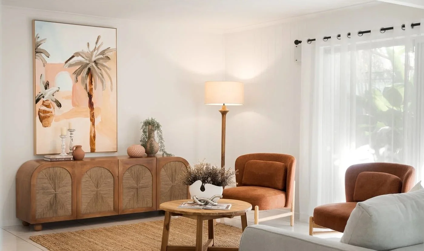 Living room with beige walls, a large abstract painting of palm trees, wooden sideboard, potted plants, coral-colored chairs, a white rug, and a window with sheer curtains.