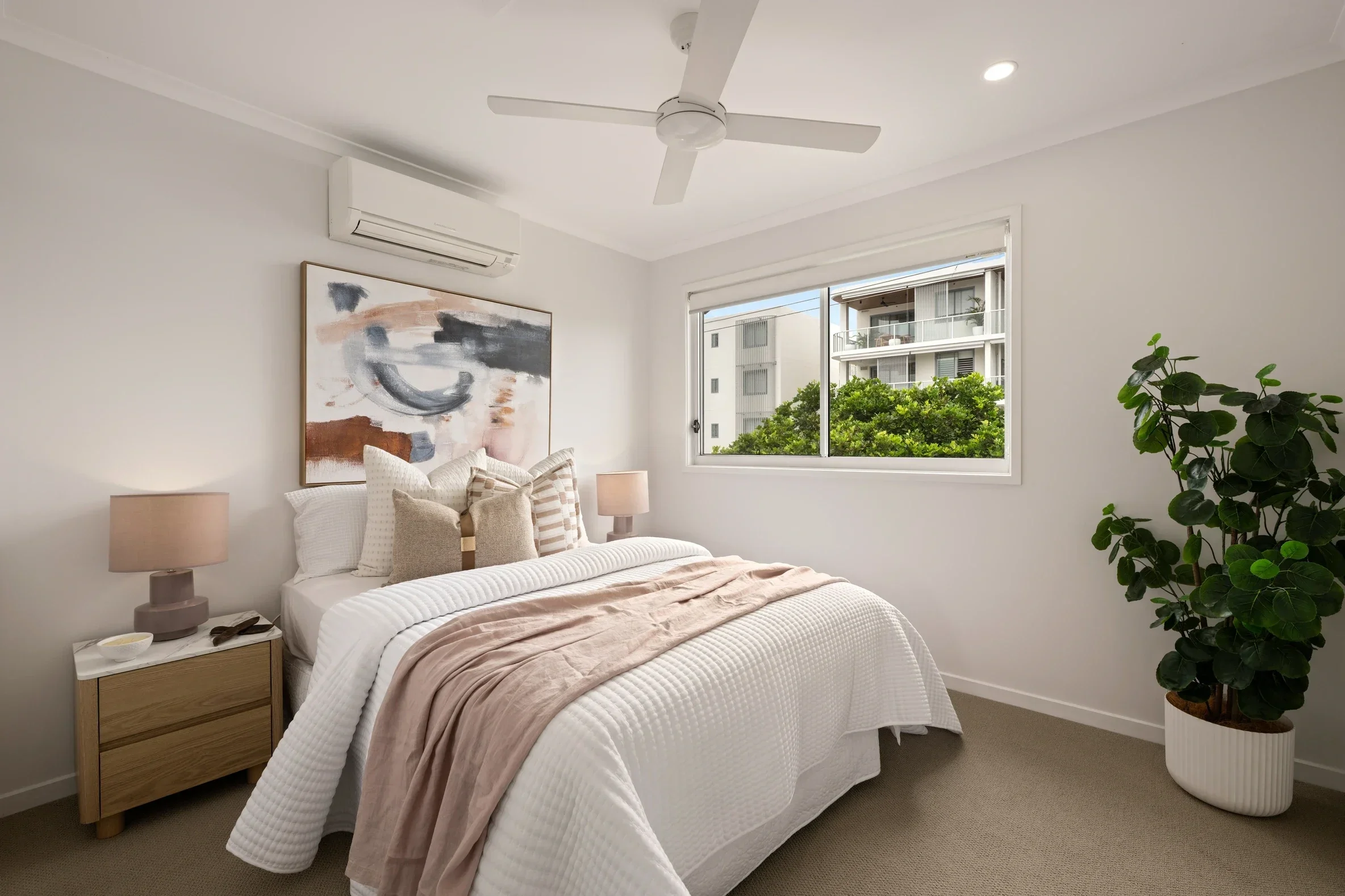 A bedroom with a white bedspread, beige blanket, and decorative pillows, wooden nightstands with beige lamps, a large abstract painting on the wall, a window with a view of a neighboring building and green trees, a potted plant in the corner, a ceiling fan, an air conditioning unit, and a recessed ceiling light.