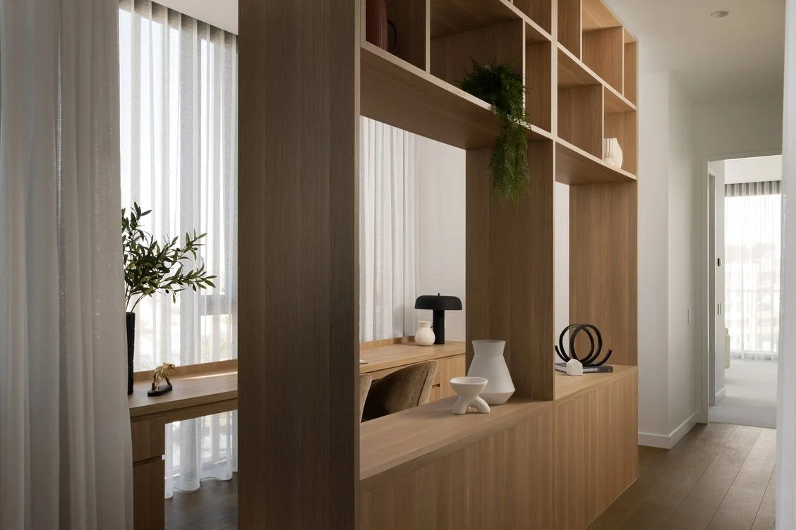 Interior view of a modern home with a wooden room divider, decorative vases, and a workspace with a chair and lamp, with sheer curtains and sunlight in the background.