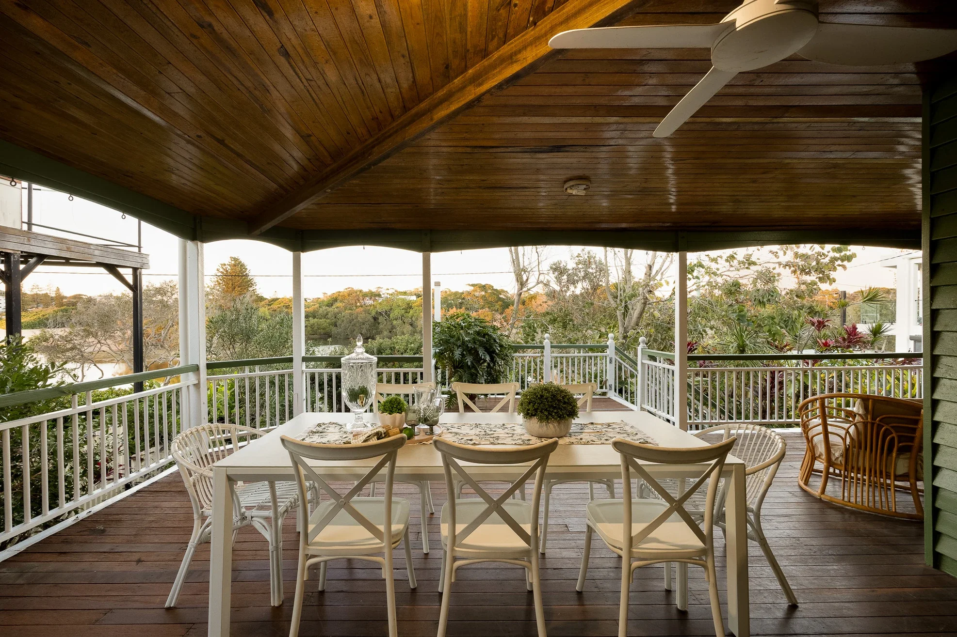 A covered outdoor patio with a wooden ceiling and white railing. There is a large dining table with chairs, a few plants, and decorative items, overlooking a garden with trees and water in the background.
