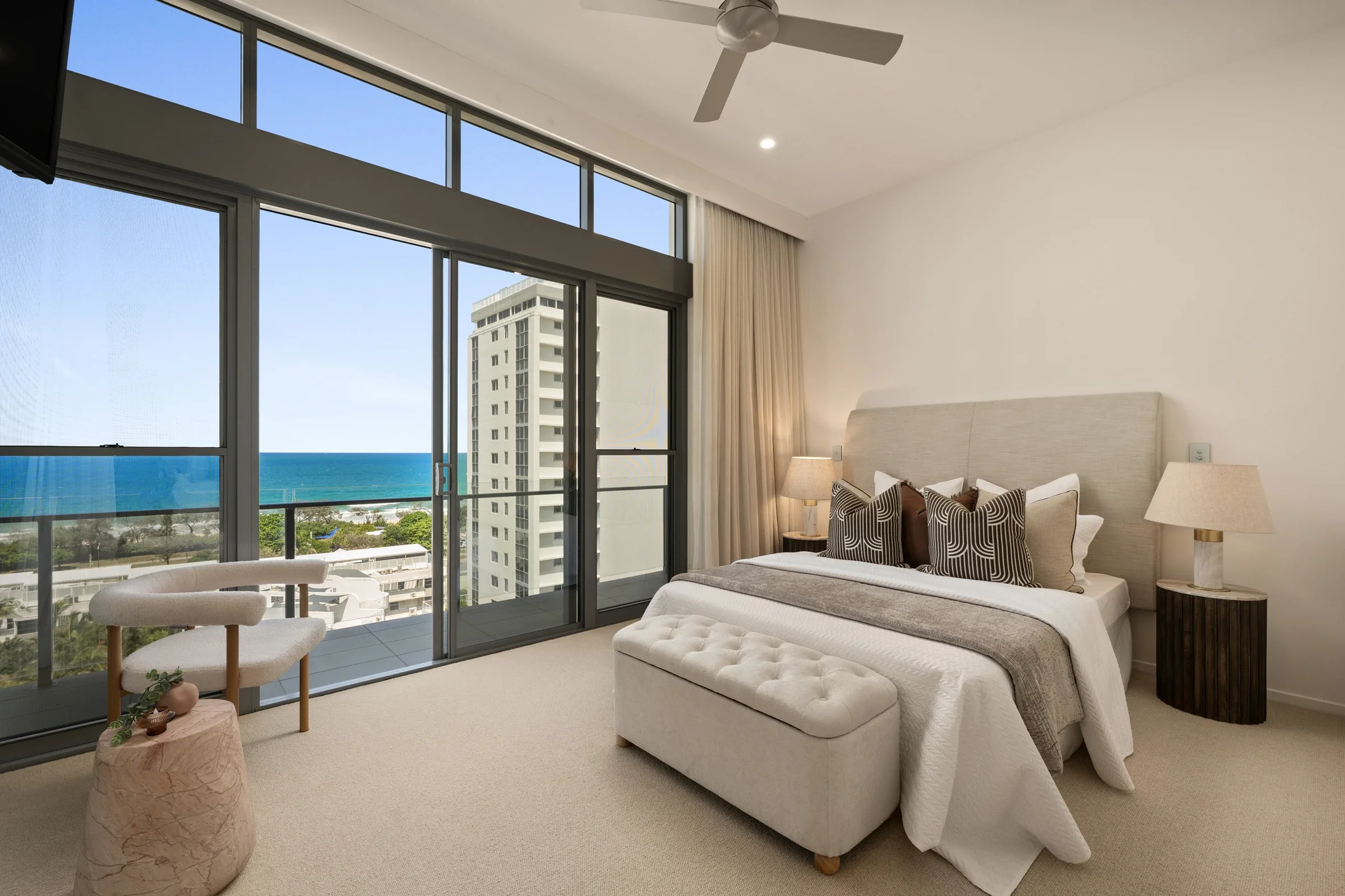 Interior of a modern bedroom with a large window overlooking the ocean and city buildings, a bed with decorative pillows, a bedside lamp, a stool with a potted plant, and a ceiling fan.
