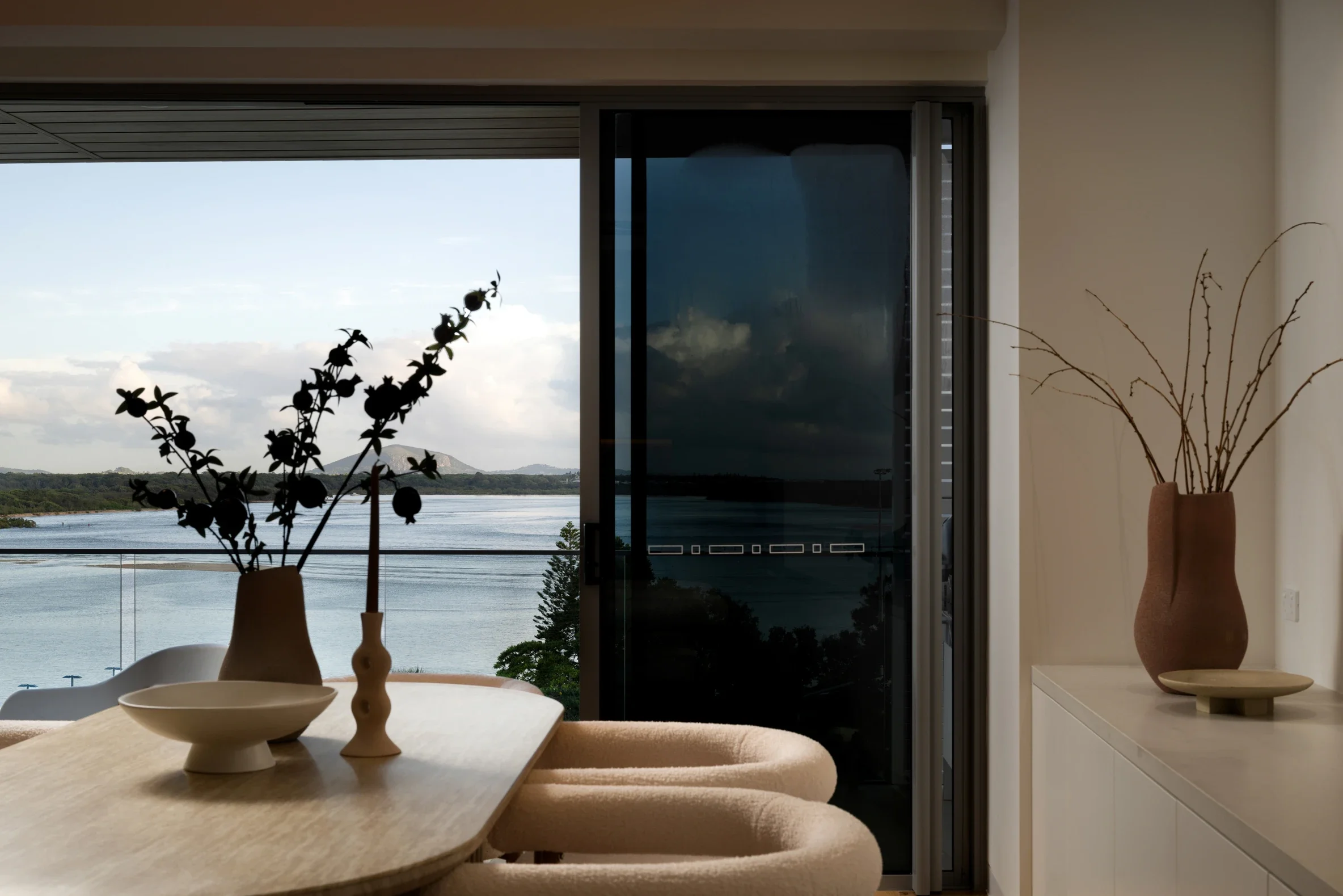 A dining room with a wooden table, beige chairs, and decorative vases, overlooking a body of water and mountains through a large sliding glass door.