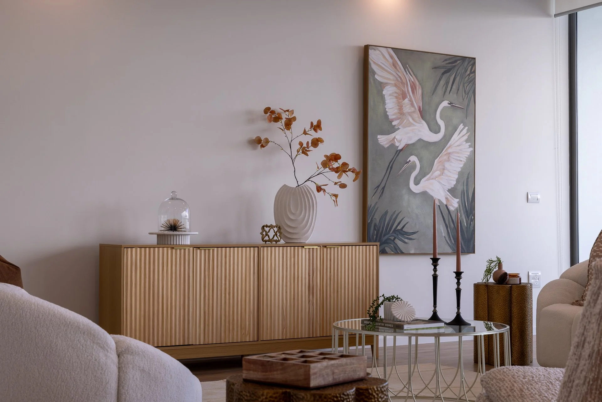 Modern living room with beige sofa, wooden sideboard, large painting of white herons, decorative vases, and candles.