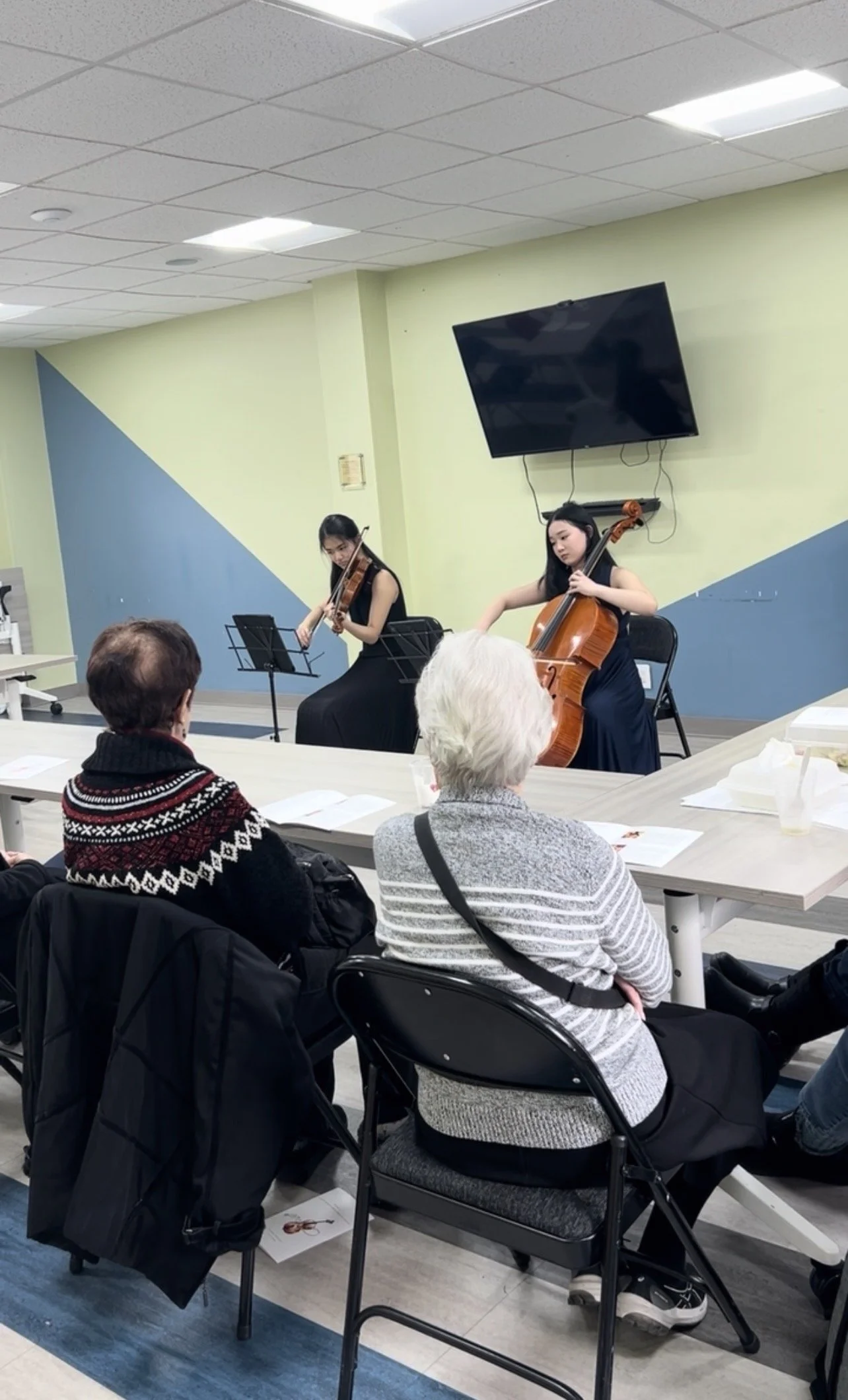 Two women playing violin and cello during a live performance for an audience in a room with pastel green and blue walls, a large TV screen mounted on the wall, and elderly audience members seated at tables, some with papers and drinks.