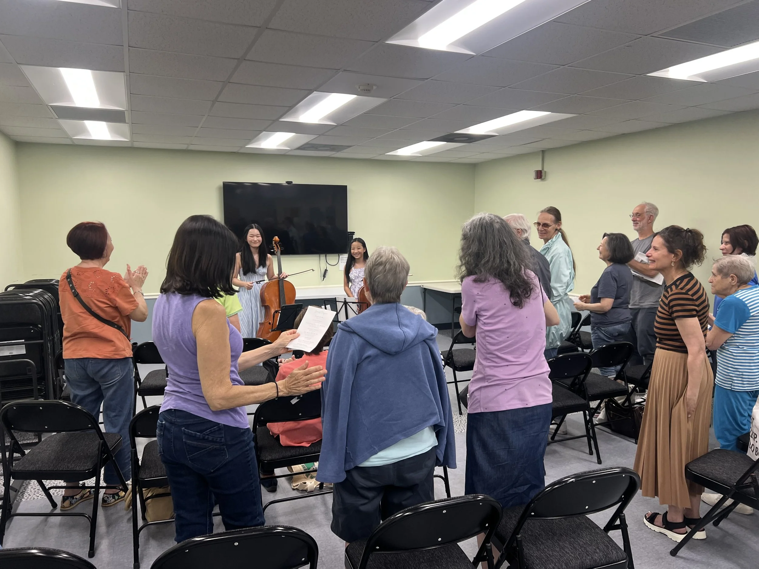 A group of people in a room listening to two young girls playing violins at the front. Some people are smiling and clapping, and the room has a large television on the wall.