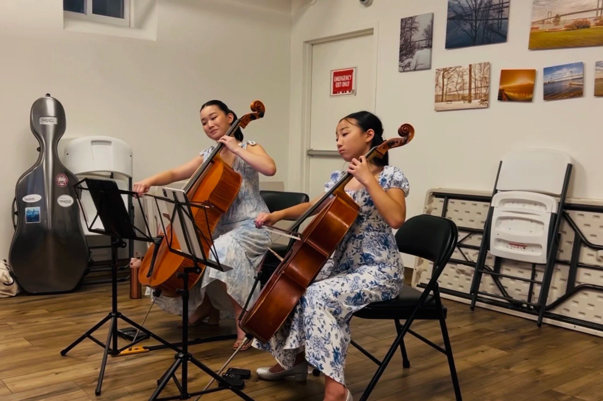 Two young girls in pastel dresses are playing cellos during a performance or practice session in a room decorated with landscape photographs on the wall. There are empty chairs and a guitar in a case nearby.