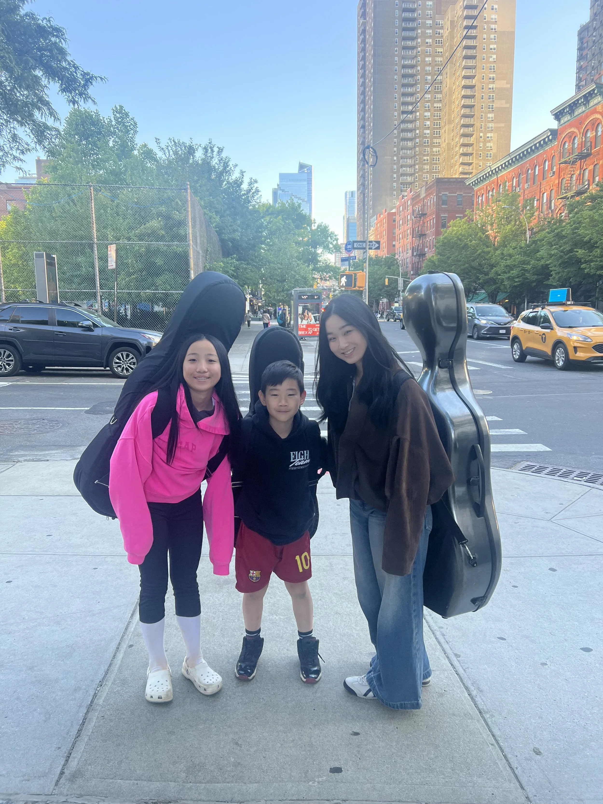 Three children and an adult standing on a city sidewalk, with tall buildings, trees, cars, and a yellow taxi in the background.