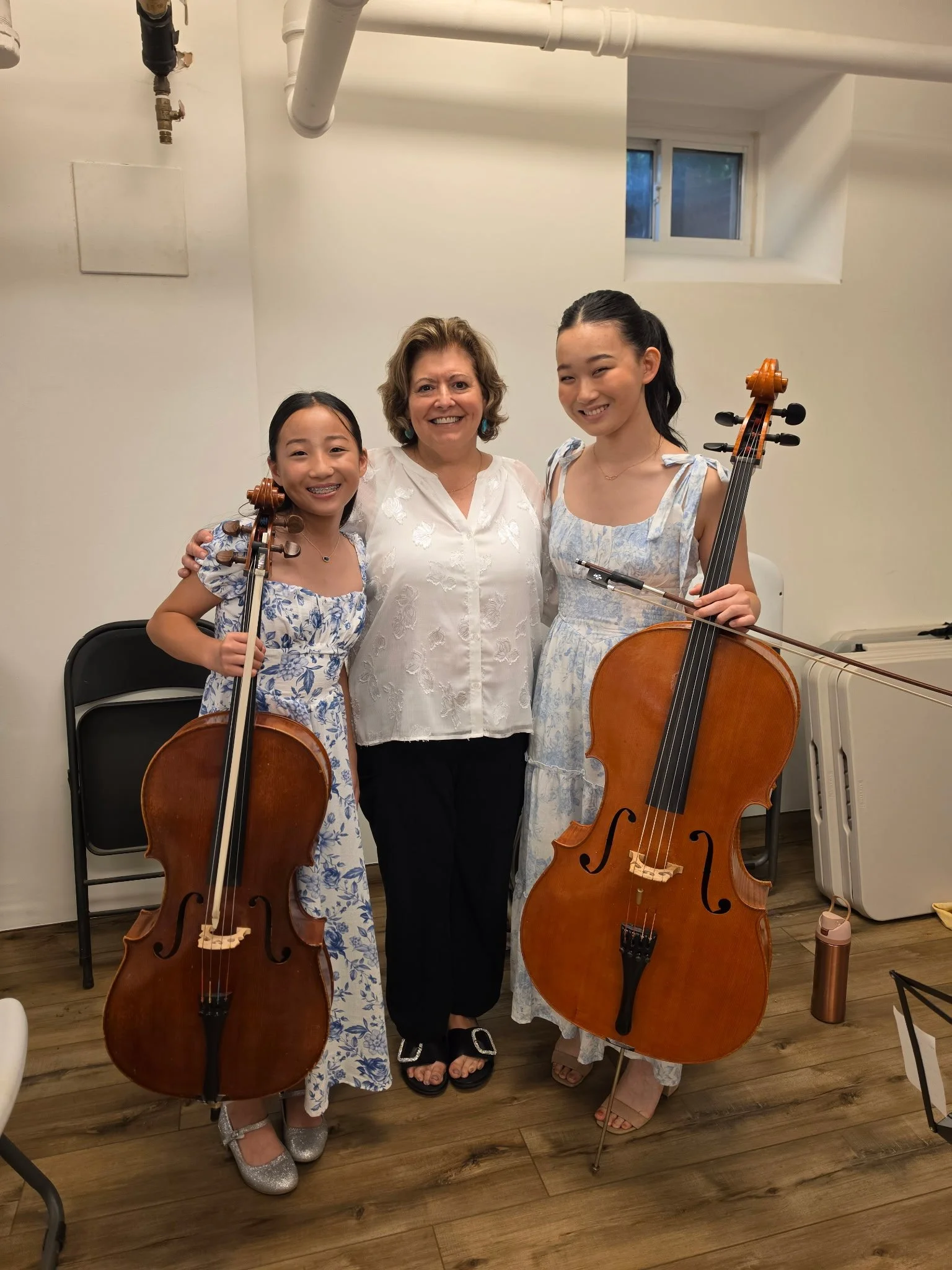 Two young girls holding cellos, standing with a smiling woman in a white blouse, in a room with wooden floors and a window. All are smiling at the camera.