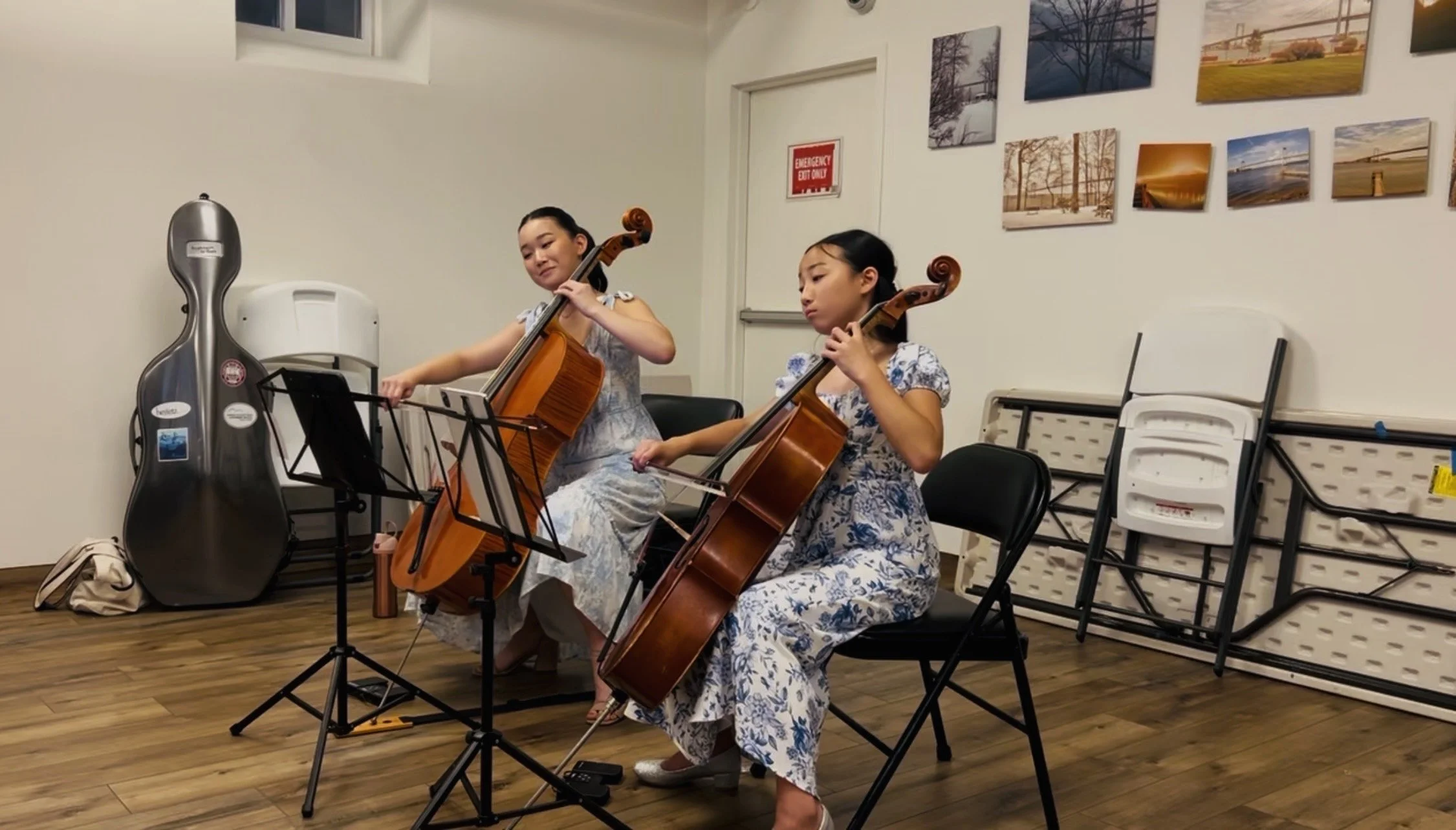 Two young girls are sitting on black chairs, playing cellos in a room with wooden floors and white walls. One girl is adjusting her music stand, while the other is focusing on her playing. There is a guitar case and a beige bag on the floor to the left. The wall behind features a small window and framed photographs of outdoor scenes. Folded chairs are stacked against the wall on the right.