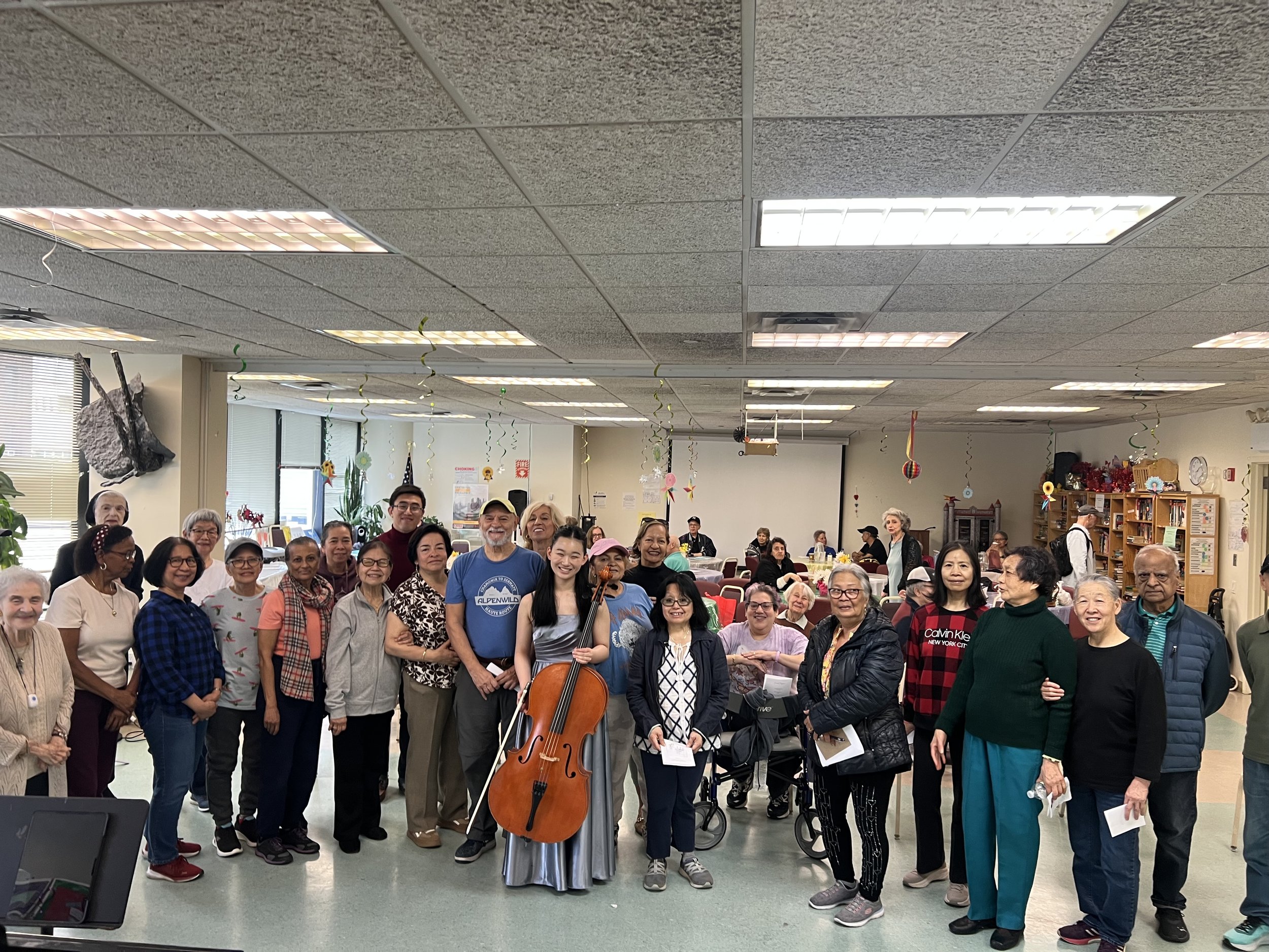 A diverse group of people gathered in a room, some standing and some sitting, with a musician holding a cello in the center, all smiling at the camera.