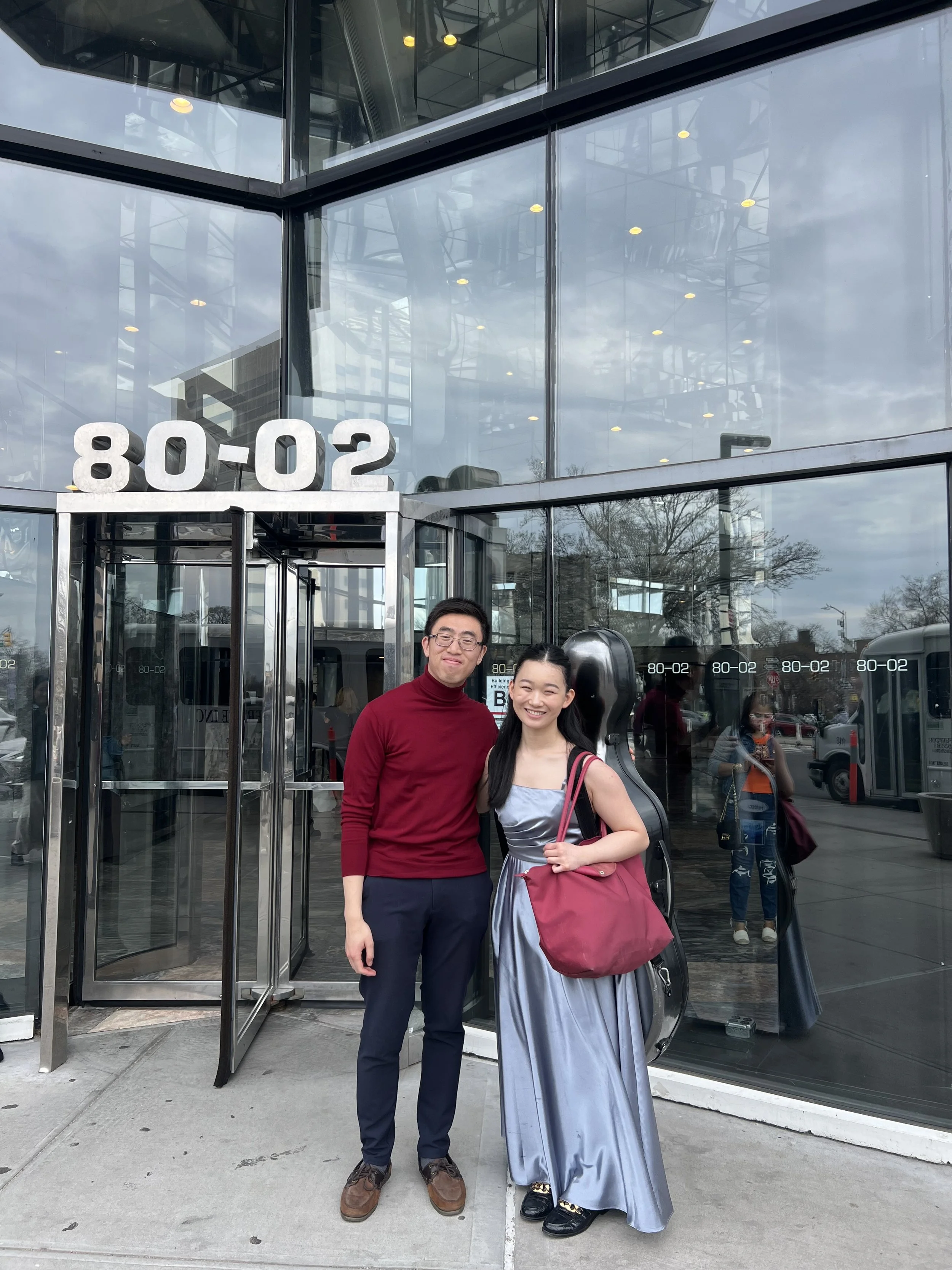 A man wearing a red turtleneck and a woman in a gray dress with a maroon bag, standing together smiling outside a modern glass building with the address 80-02 above the entrance.