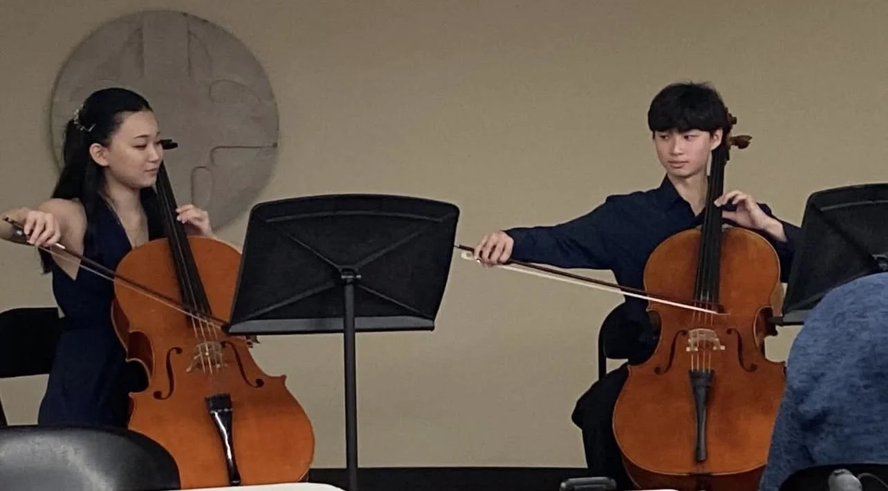 Two young musicians, a girl and a boy, playing cellos during a performance in a room with a wall clock in the background.