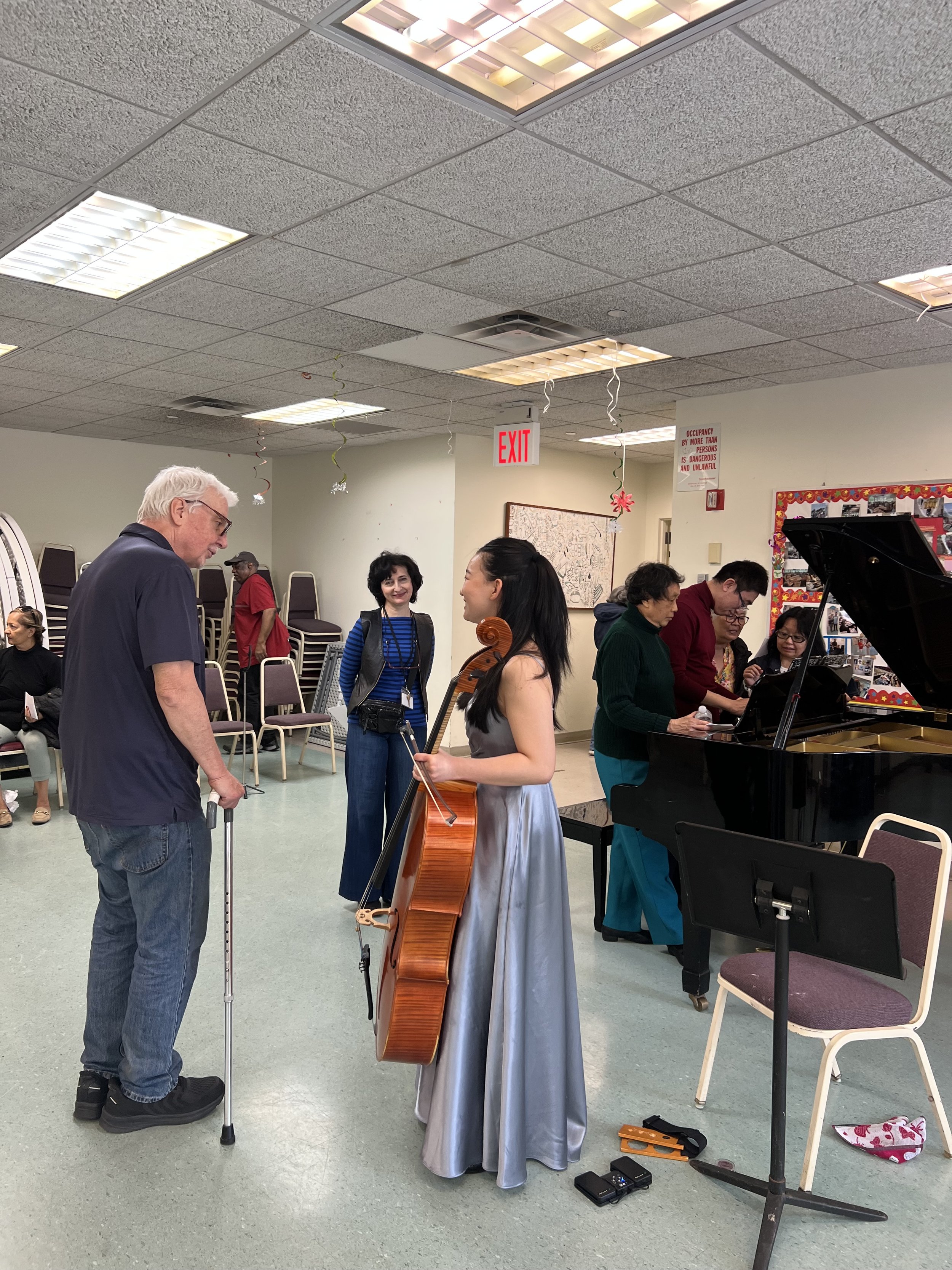 A young woman playing the cello, engaging with an elderly man with a cane, in a room decorated for a celebration. Several people are gathered, some playing instruments and others watching.