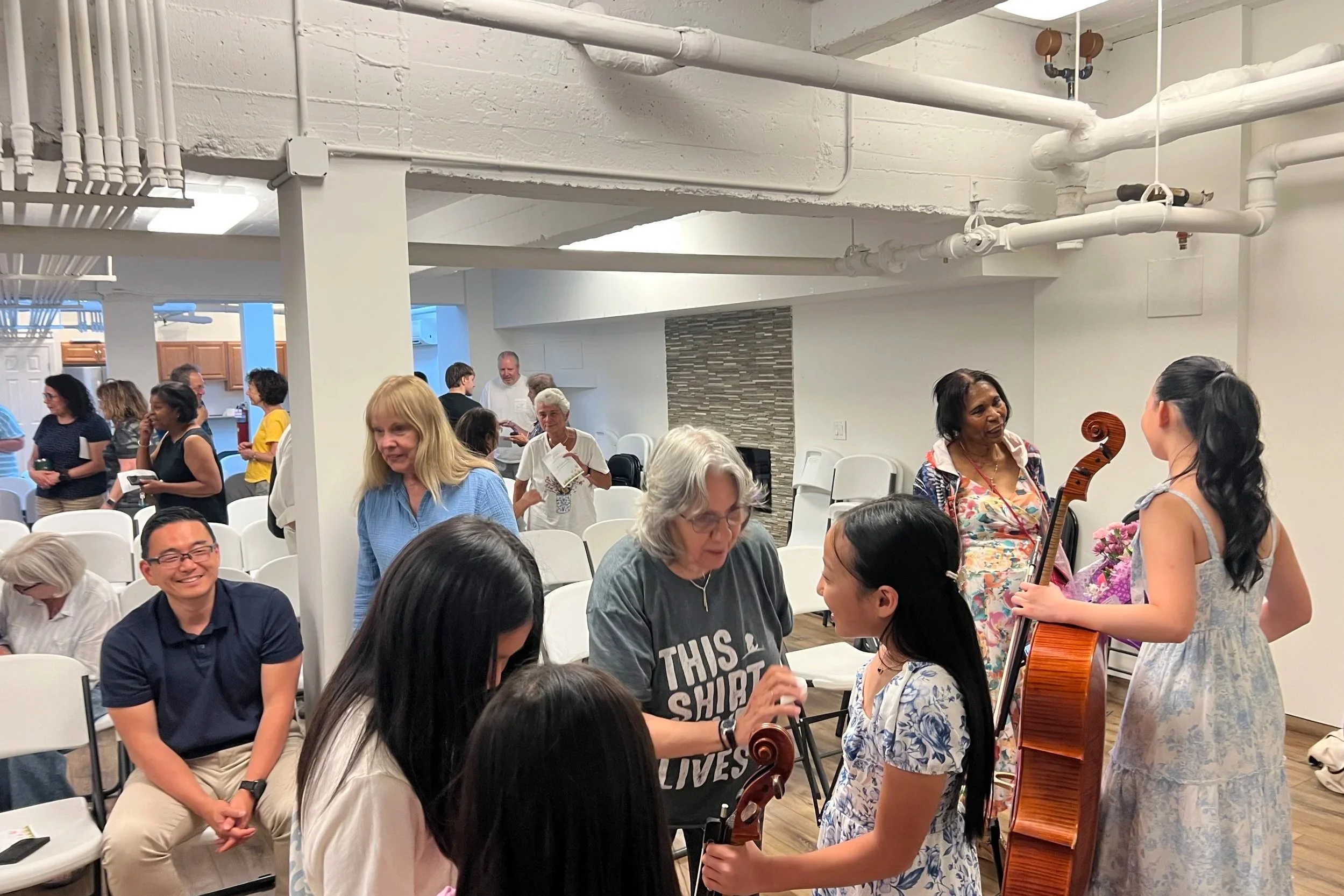 People gathered in a room with chairs, some holding musical instruments like violins, engaging in conversation and music activities.
