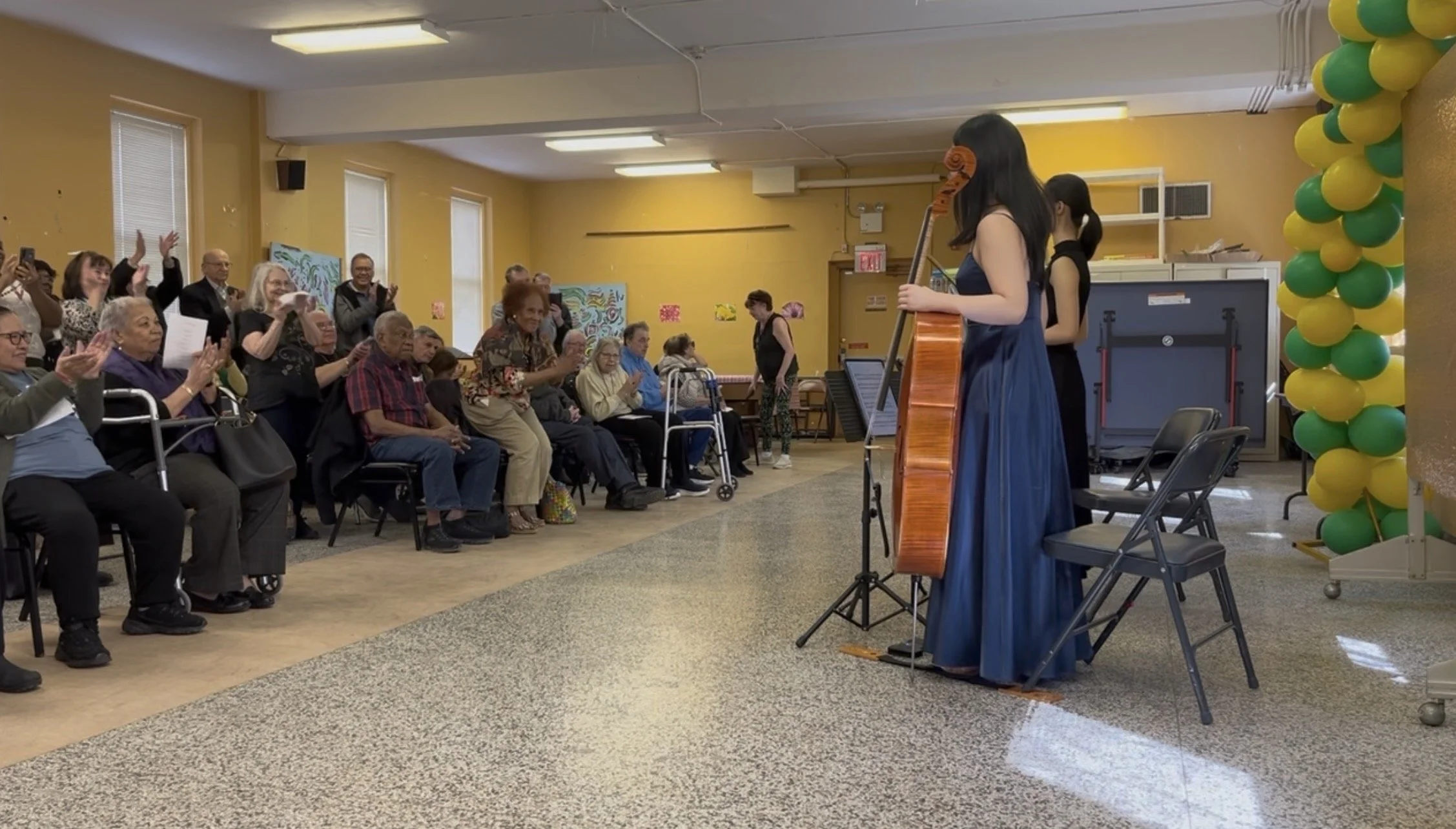 A musical performance at a senior center, featuring a woman in a blue dress playing an acoustic guitar while an audience of seniors claps and smiles.