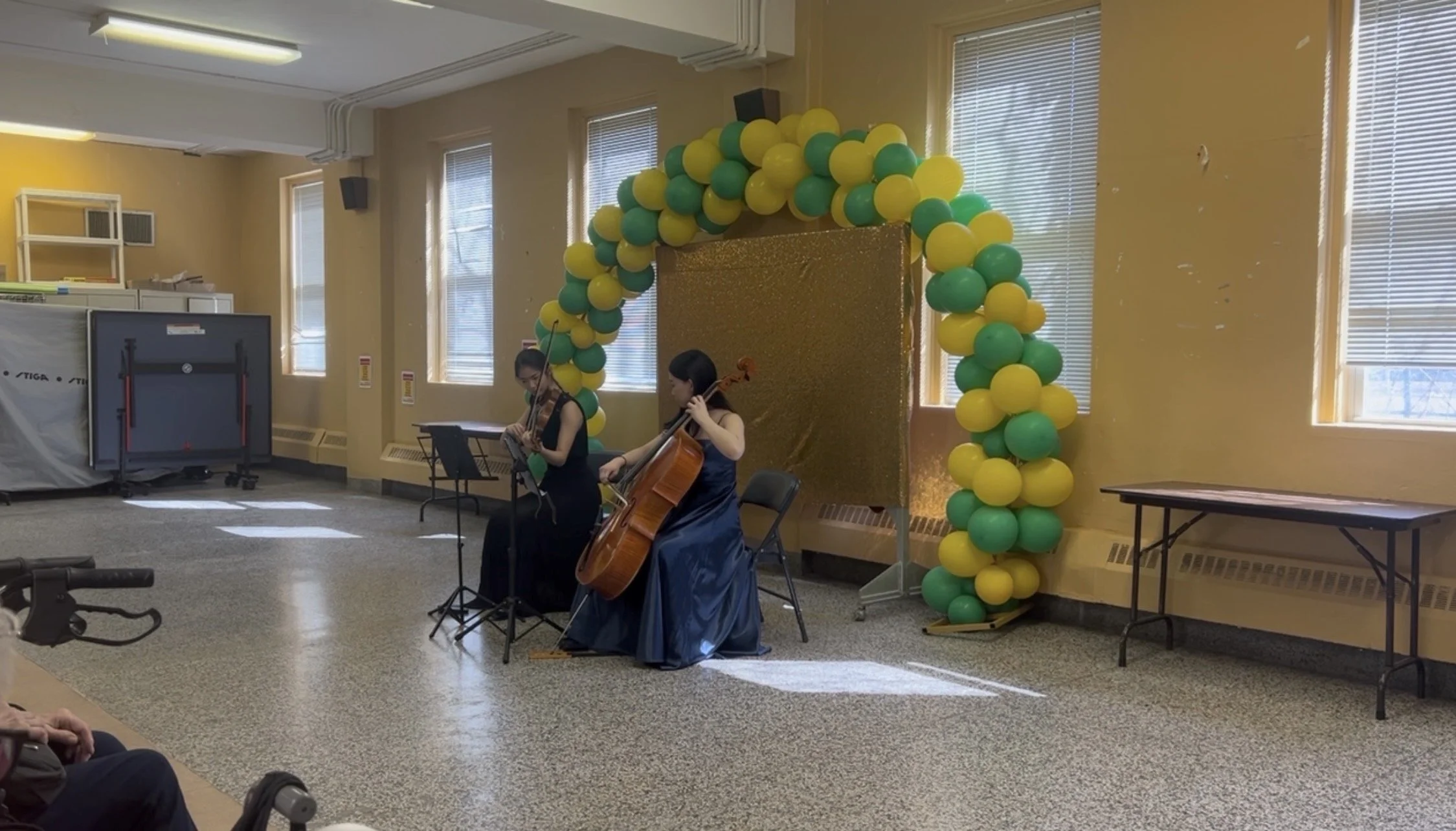 Two women playing string instruments, one with a violin and the other with a cello, performing in front of a golden backdrop decorated with yellow and green balloons, in a room with four windows and a small audience.