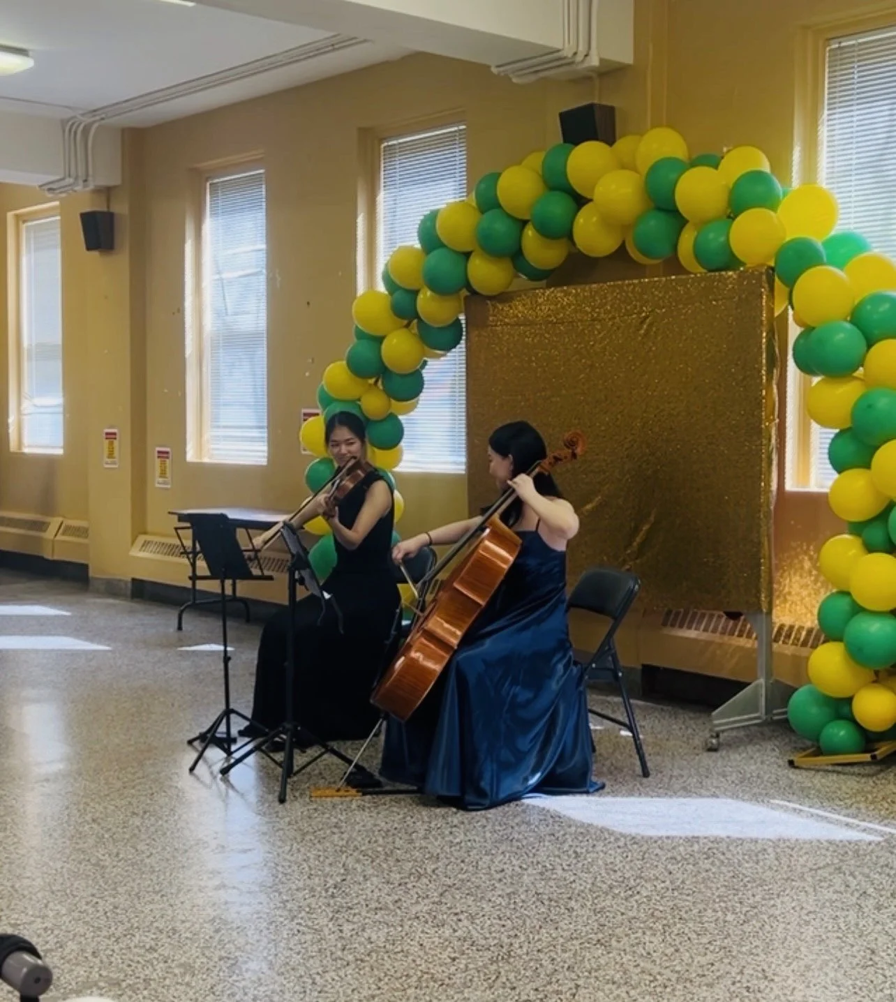 Two women in formal dresses performing a musical duet with a violin and a cello in front of a gold backdrop and balloon arch, inside a room with yellow walls and windows.