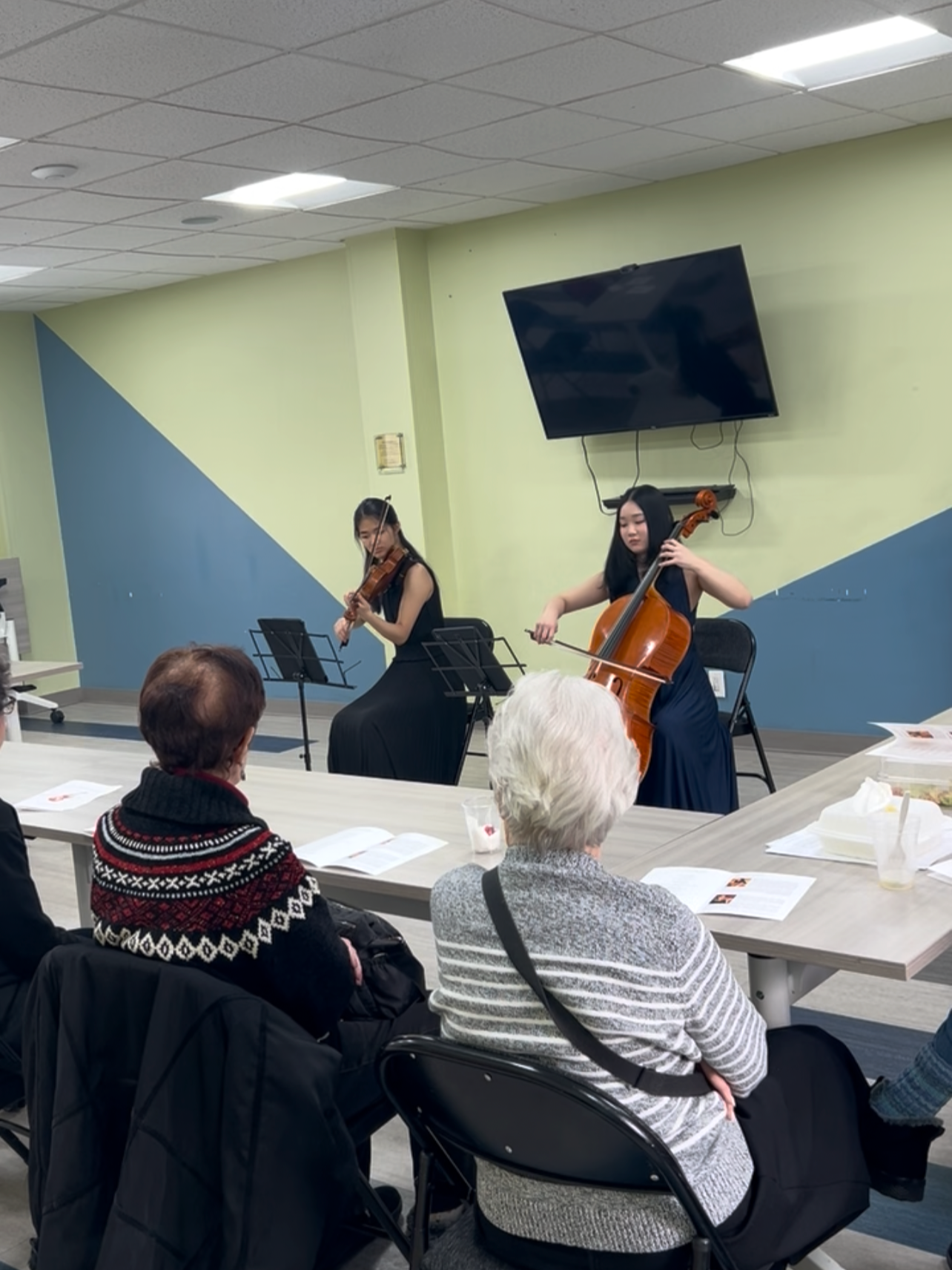 Two women playing string instruments, a violin and a cello, performing for an audience in a room with pastel-colored walls and a television mounted on the wall.