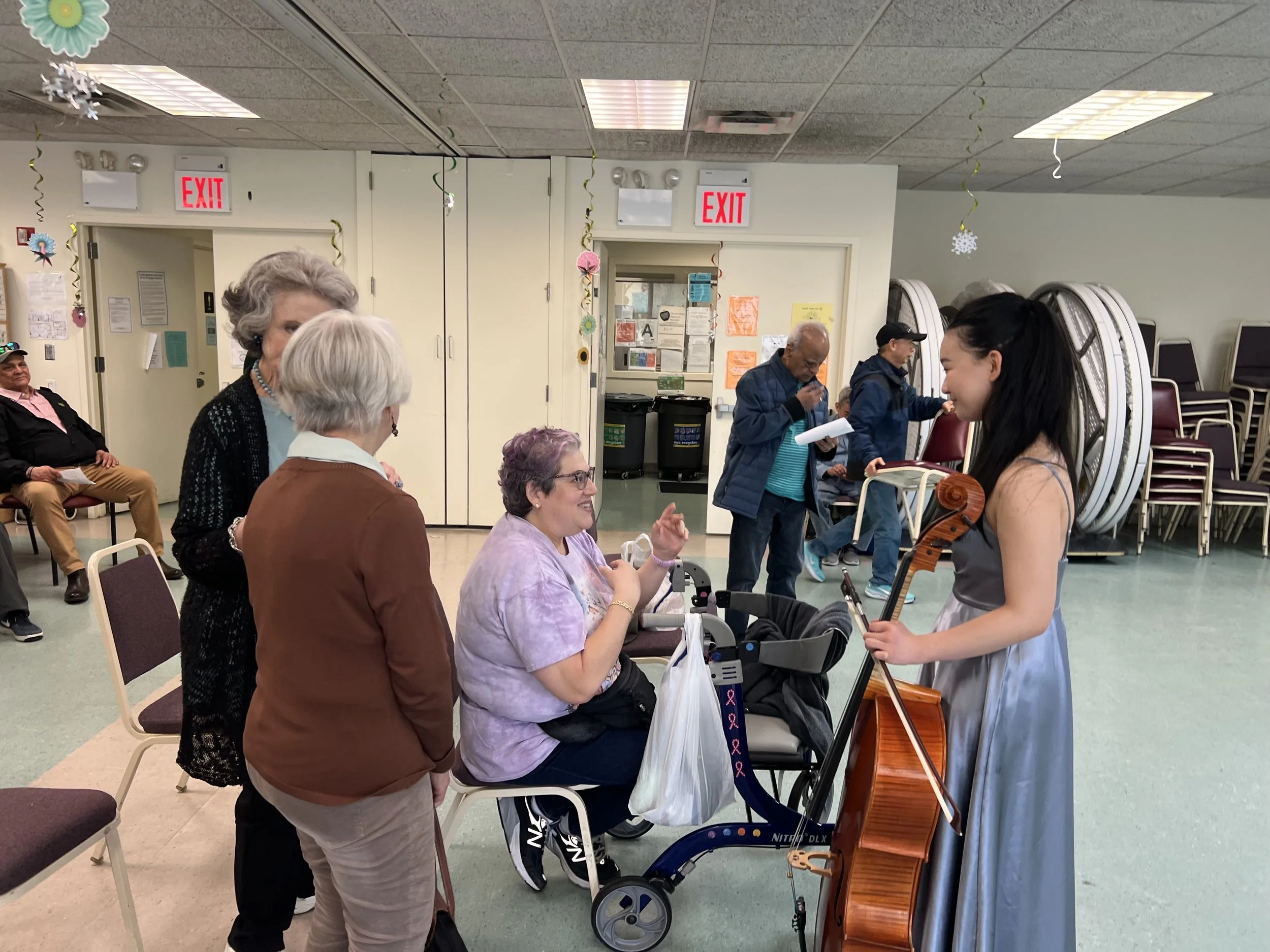 Group of elderly people and a young woman with a violin in a room with stacked chairs and Christmas decorations.