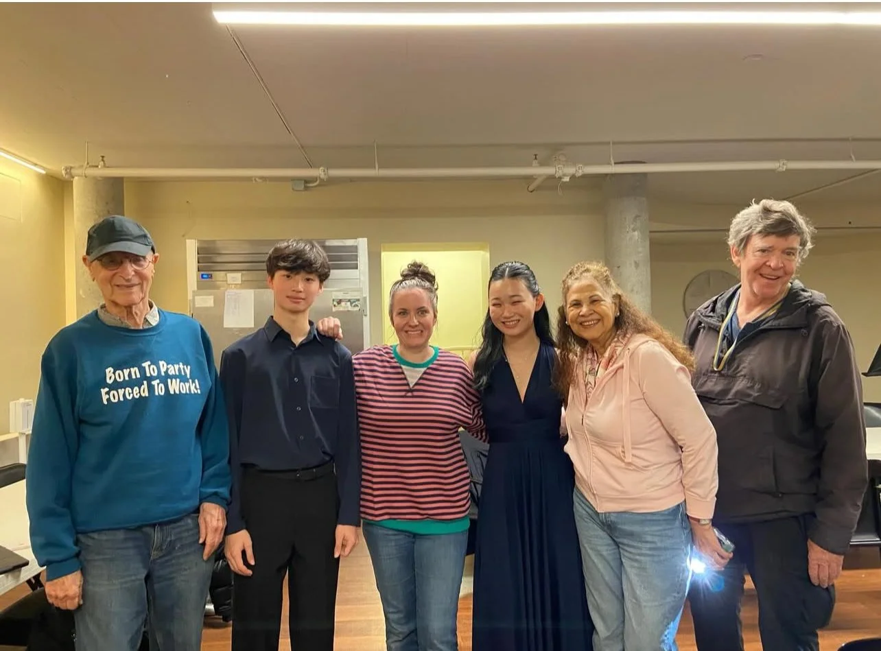 Six people standing together in an indoor setting, smiling at the camera. The group includes an elderly man with glasses and a cap, a young man in a dark shirt, three women in casual clothing, and another man in a dark jacket. The background shows a yellow wall, concrete pillar, and some office equipment.