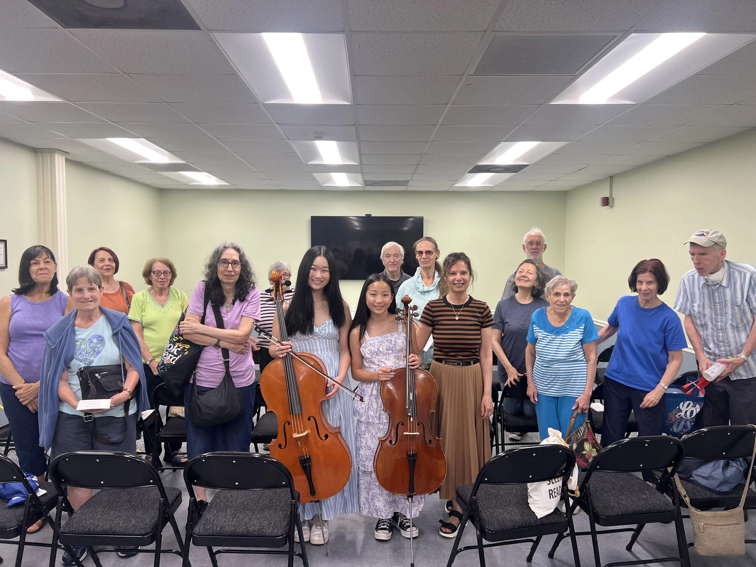 A diverse group of people of different ages and ethnicities gathered in a room with green walls and a ceiling with fluorescent lights, some holding musical instruments like violins and cellos, smiling and looking at the camera.