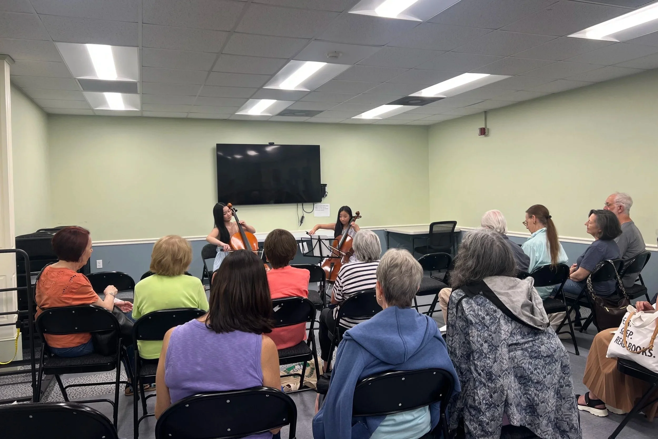 A group of people sitting in chairs watching musicians play violins in a room with a large television on the wall and bright fluorescent lighting.
