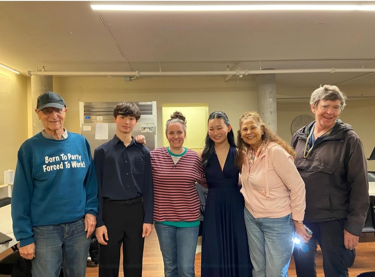 Six people standing indoors, smiling, posing together for a group photo in a room with pipes on the ceiling.