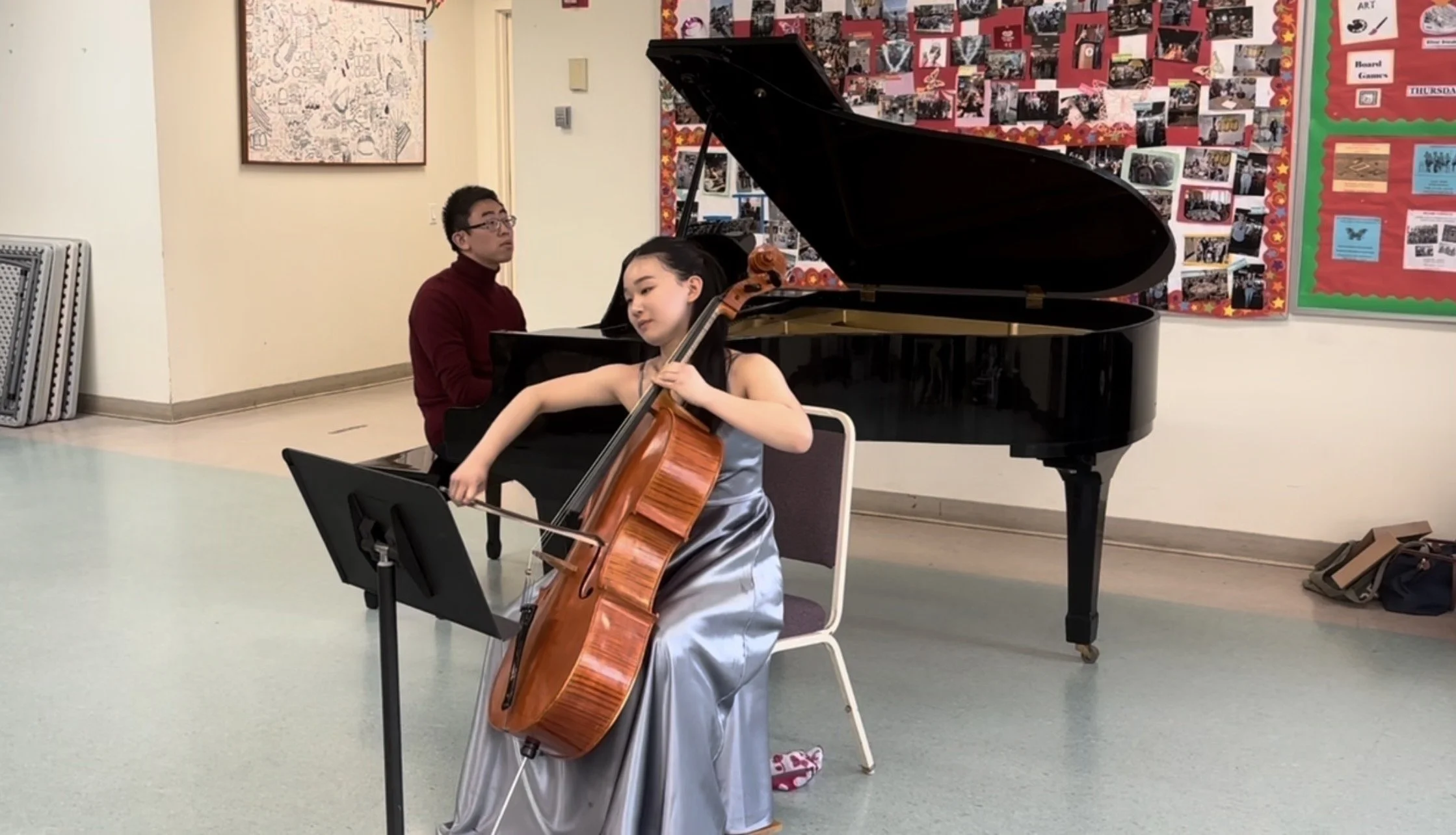 A young girl in a silver dress playing the cello while a man in a burgundy turtleneck watches. They are in a school or community center with colorful bulletin boards and photos on the walls.
