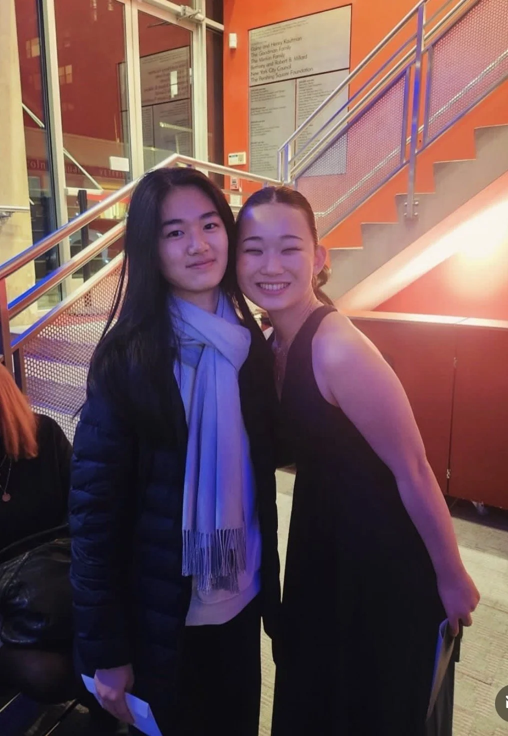 Two women standing close together, smiling, in an indoor setting with orange walls and a staircase in the background.