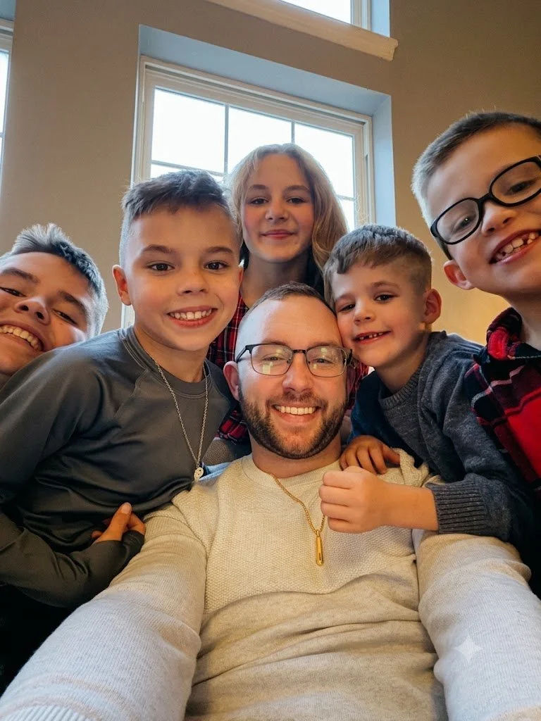 A smiling man with glasses surrounded by five smiling children in a cozy indoor setting with a window in the background.
