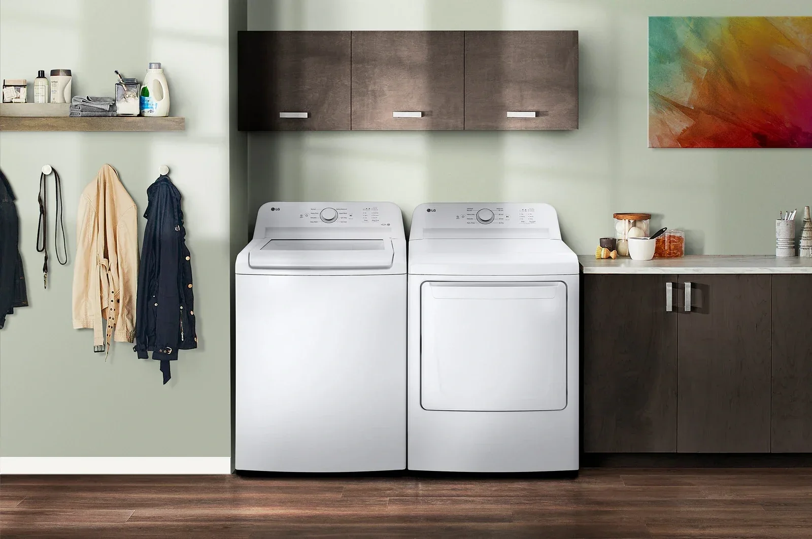 Laundry room with a side-by-side washer and dryer, dark wood cabinets, a countertop with containers and utensils, and hanging coats on the wall.
