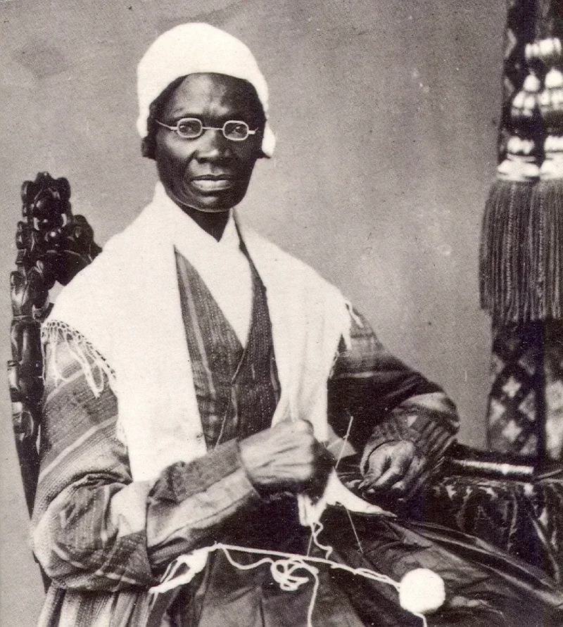 Photo of Black woman wearing a sewing cap and glasses with sewing needles in hand and a ball of yarn in her lap.