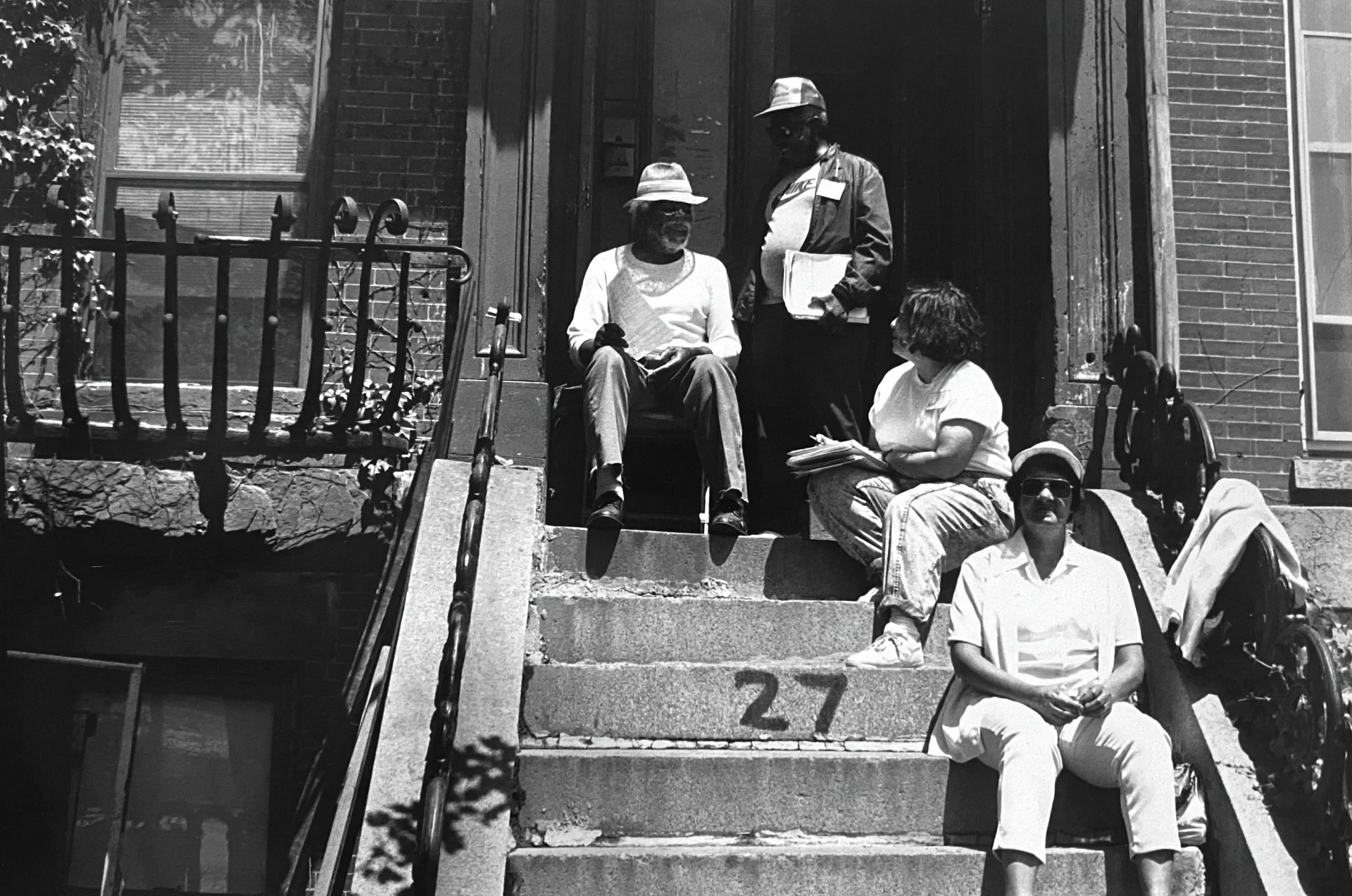 Black and white photo of organizers with flyers and clipboards  sitting on a front stoop talking to a resident.