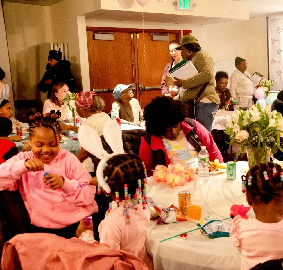 Photo of volunteers talking with ESWA members at a party with children playing in the foreground.