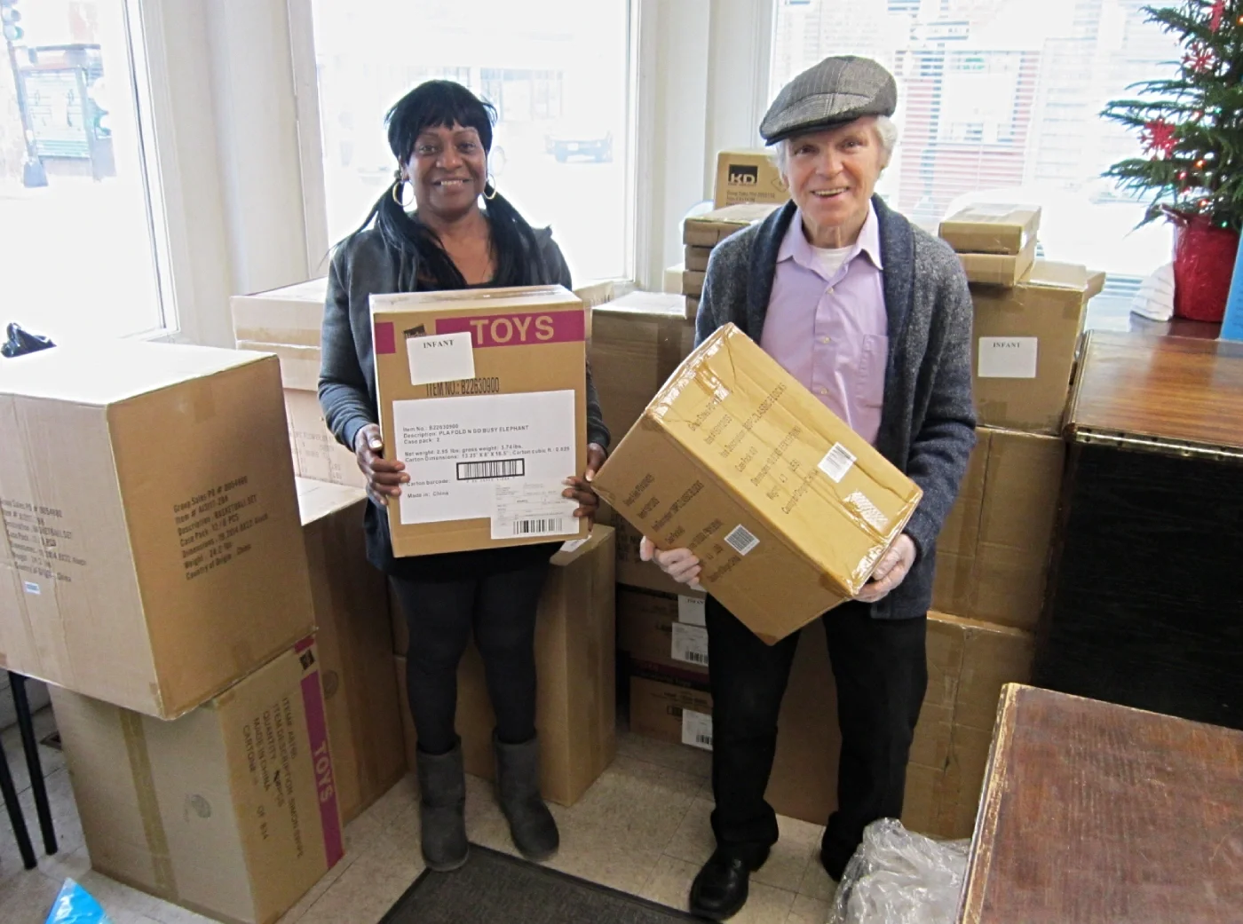 Two volunteers carrying boxes of donated toys, surrounded by stacks of other toy boxes.