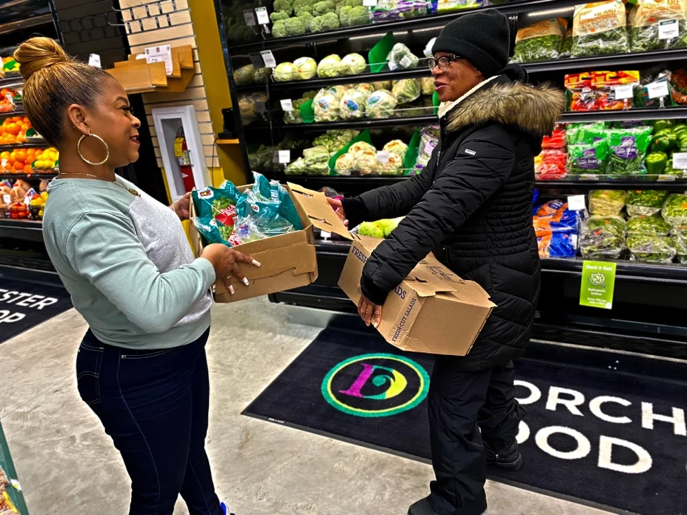 Photo of smiling grocery worker handing a box of salad greens to a smiling volunteer.