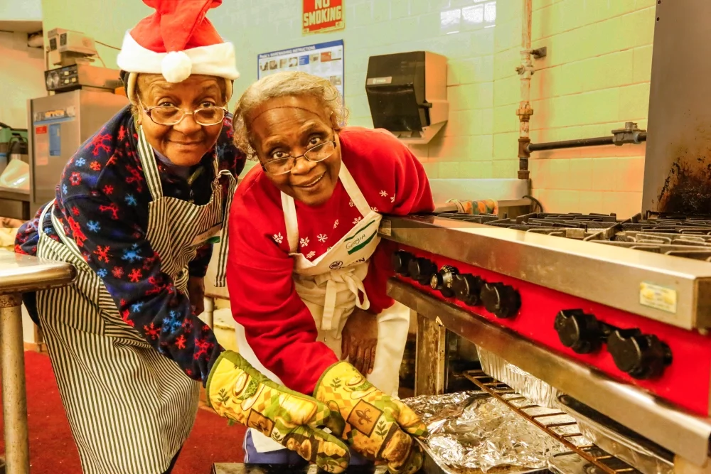 Two ESWA members both wearing festive Christmas clothes pull a baking tray out of an oven.