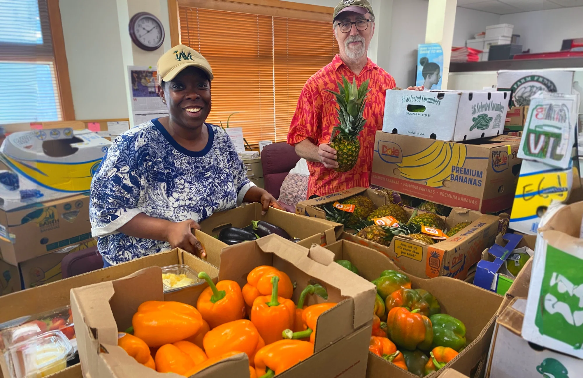 Smiling volunteers sorting produce. One volunteer holds up a pineapple, another holds a case of eggplants.