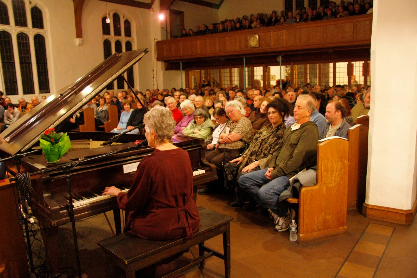 Photo of woman playing piano in front of a crowd of people sitting in church pews and on a balcony.