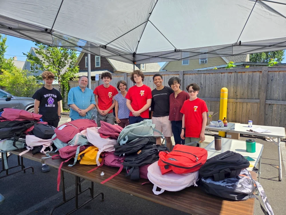 Group photo of volunteers outdoors under a tent and behind a table covered in donated backpacks.