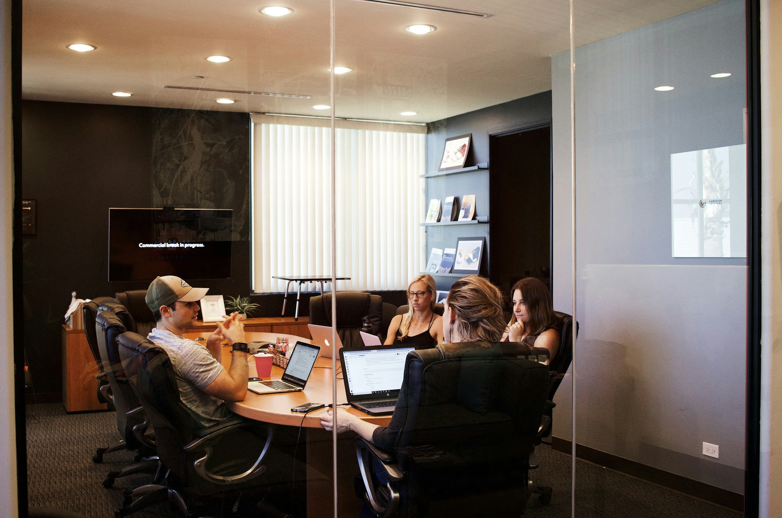 A group of people sitting around a conference table in a meeting room, working on laptops, with a black wall, bookshelves, and a window with vertical blinds in the background.