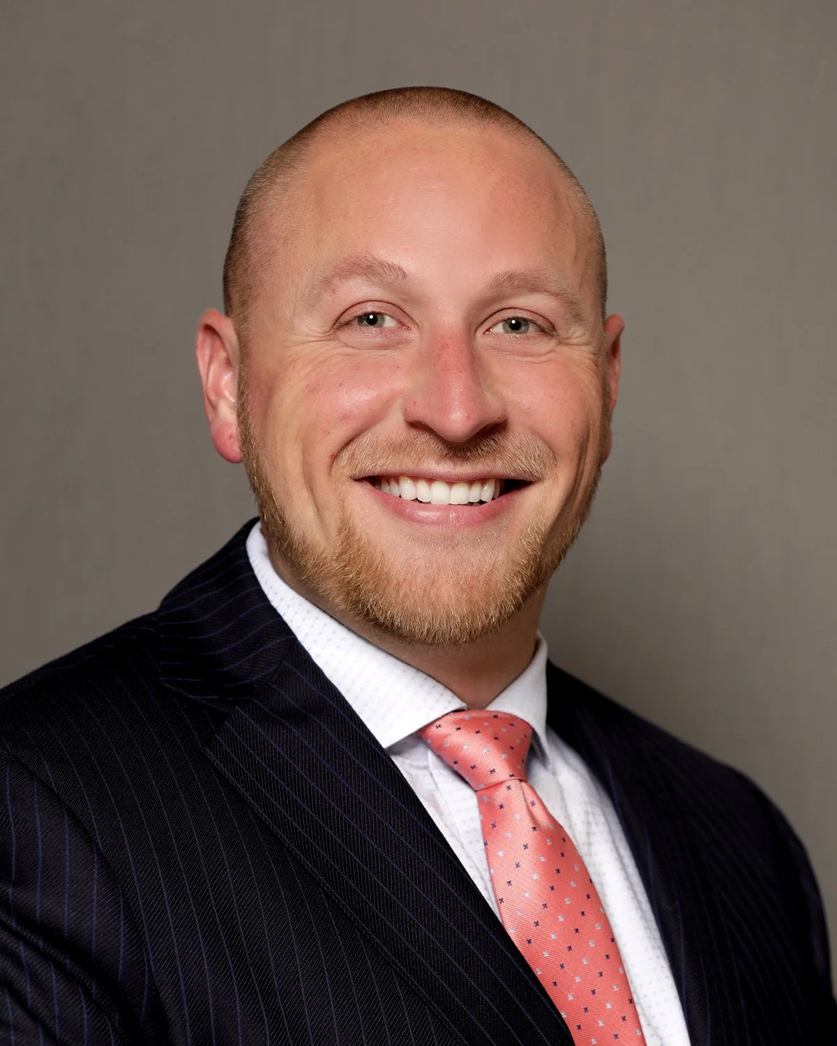 A smiling man in a dark pinstripe suit, white shirt, and pink tie with small dots, against a plain gray background.