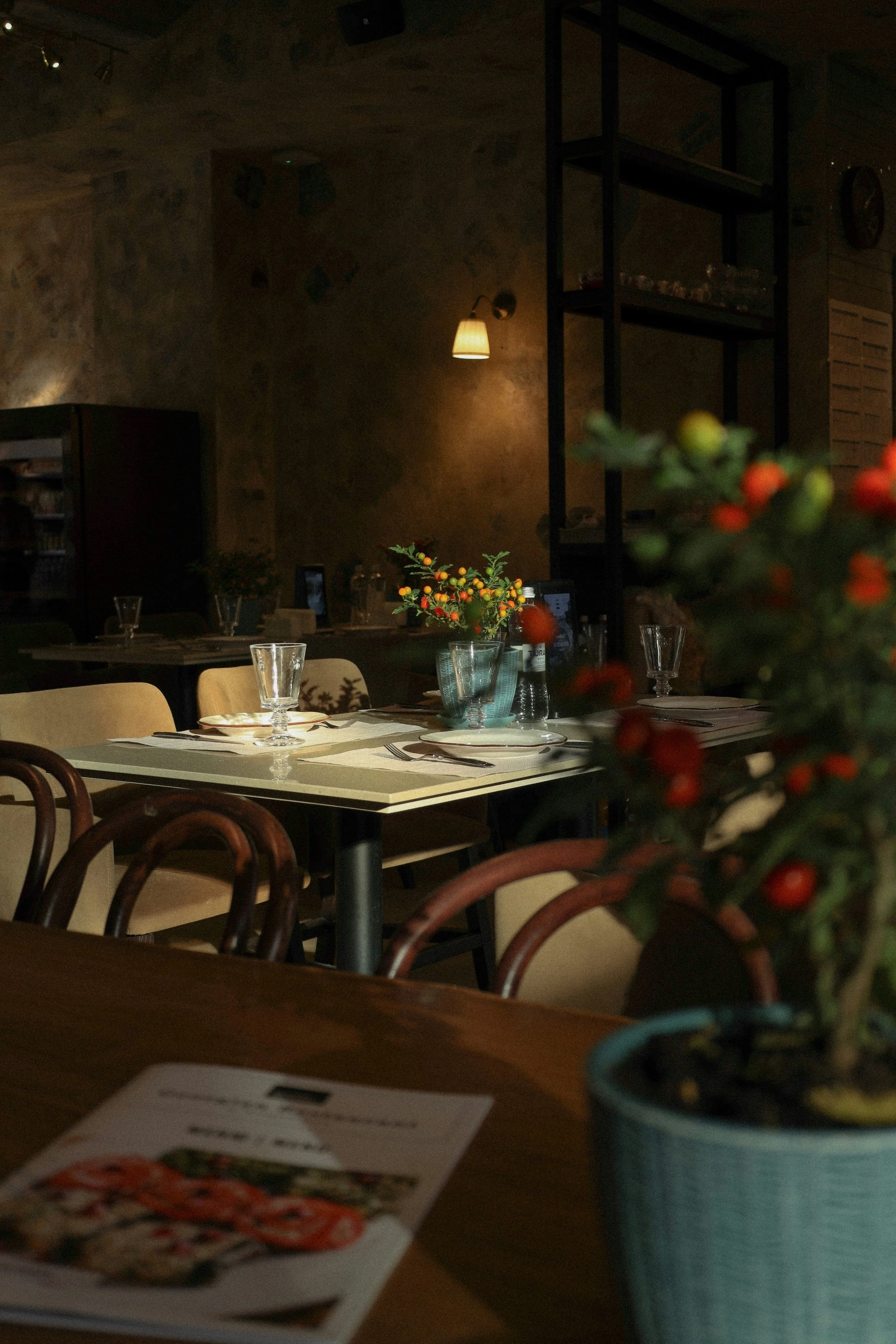 A dimly lit restaurant or café with a table set with glasses and plates, decorated with a small bouquet of colorful flowers, and surrounded by chairs. There is a tall shelving unit and a vending machine against a textured wall, and a wall-mounted lamp provides warm lighting.