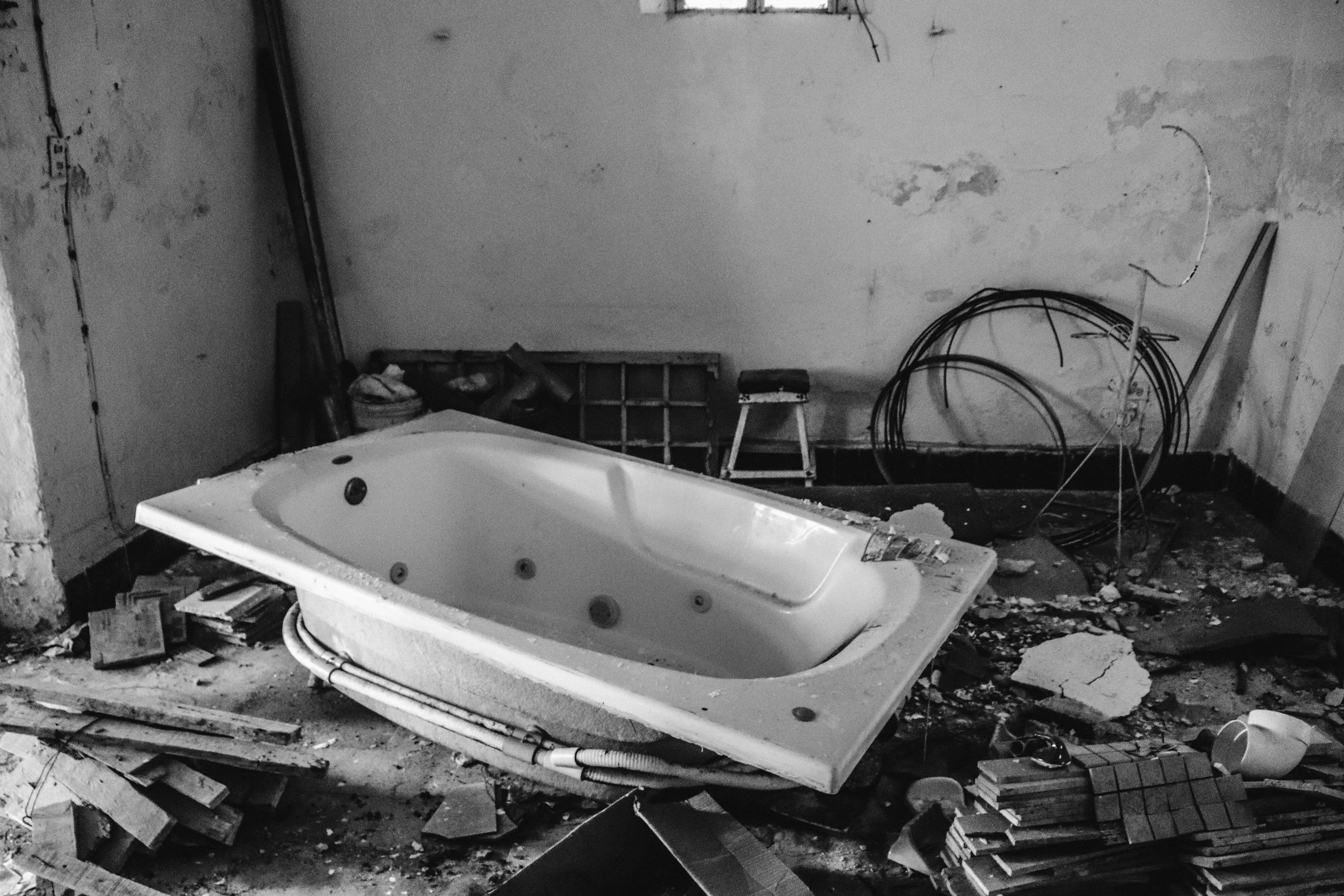 A cluttered and dusty bathroom with a large, abandoned bathtub, surrounded by construction debris, wood planks, and exposed wiring on the walls.